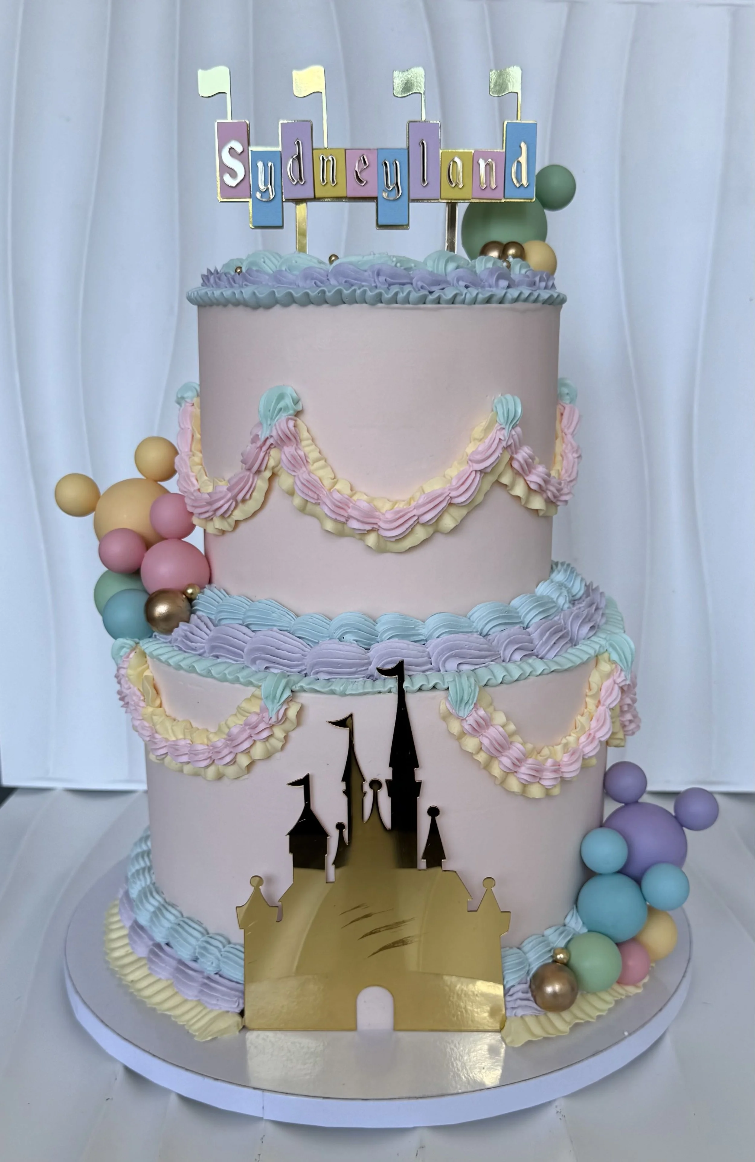 Pastel de cumpleaños de varios niveles decorado con flores de colores suaves, globos y un castillo dorado en la base, con una bandera de 'Sydneyland' en la parte superior.