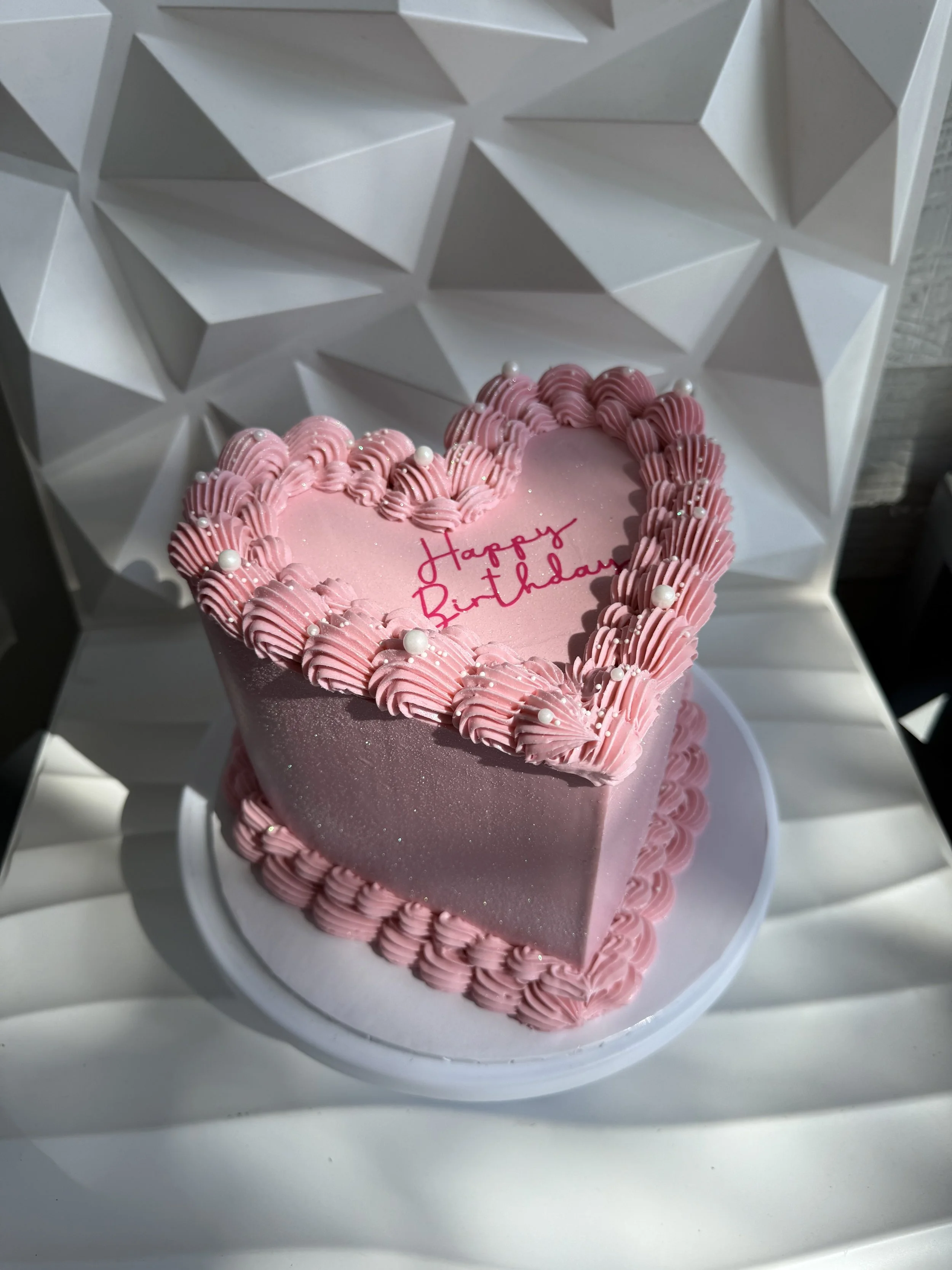 Pastel de cumpleaños en forma de corazón con decoraciones rosas y pequeñas perlas blancas, con la inscripción 'Happy Birthday' en el centro, en un plato blanco.