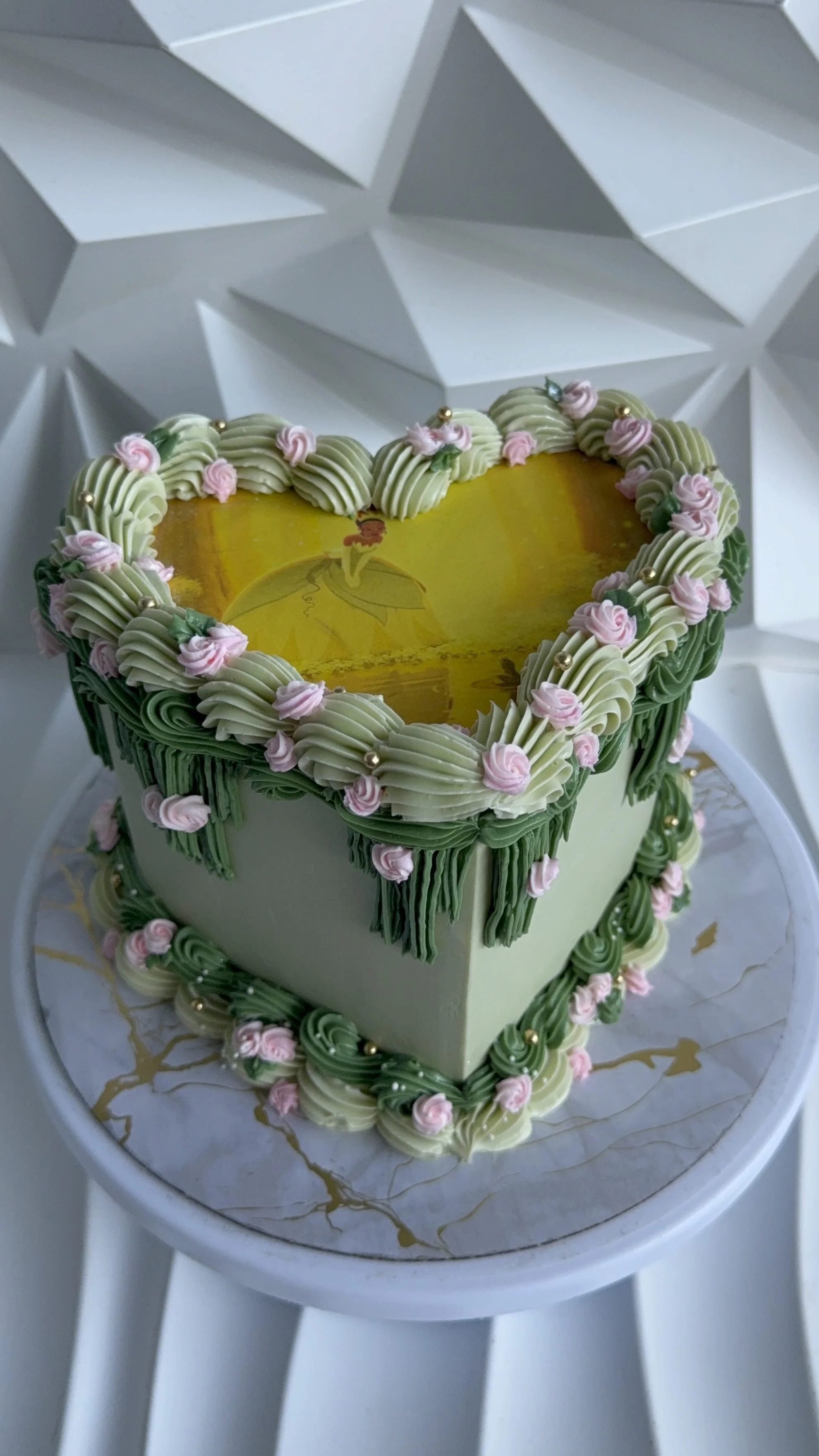Pastel de color verde con decoraciones en rosa, blanco y dorado, en forma de corazón, en un plato con dibujos dorados.