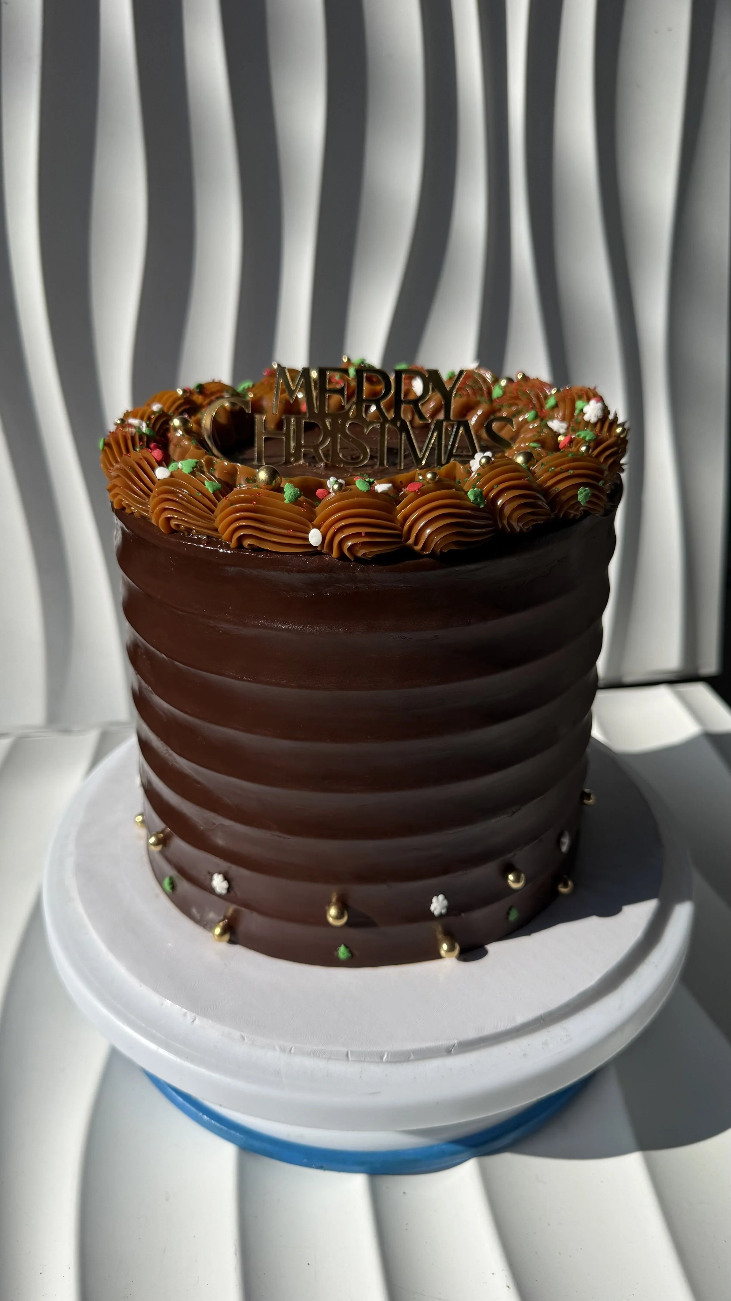 Pastel de chocolate grande decorado con glaseado de chocolate, suficientes glaseado de colores y pequeños adornos de Navidad, en una mesa blanca con fondo de cortinas blancas