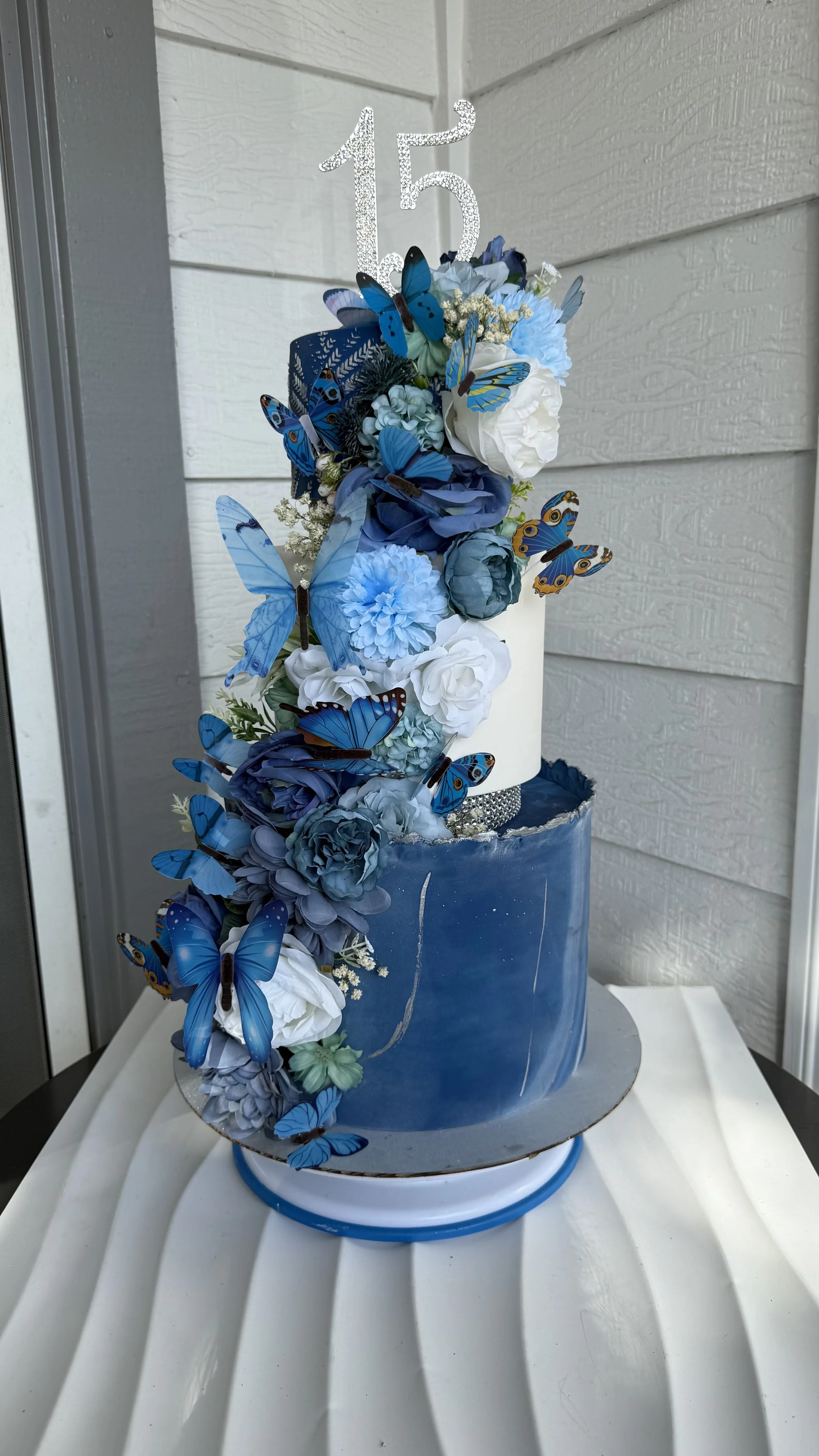 Pastel de cumpleaños azul decorado con flores blancas y azules y mariposas azules, con un topper de número 15.