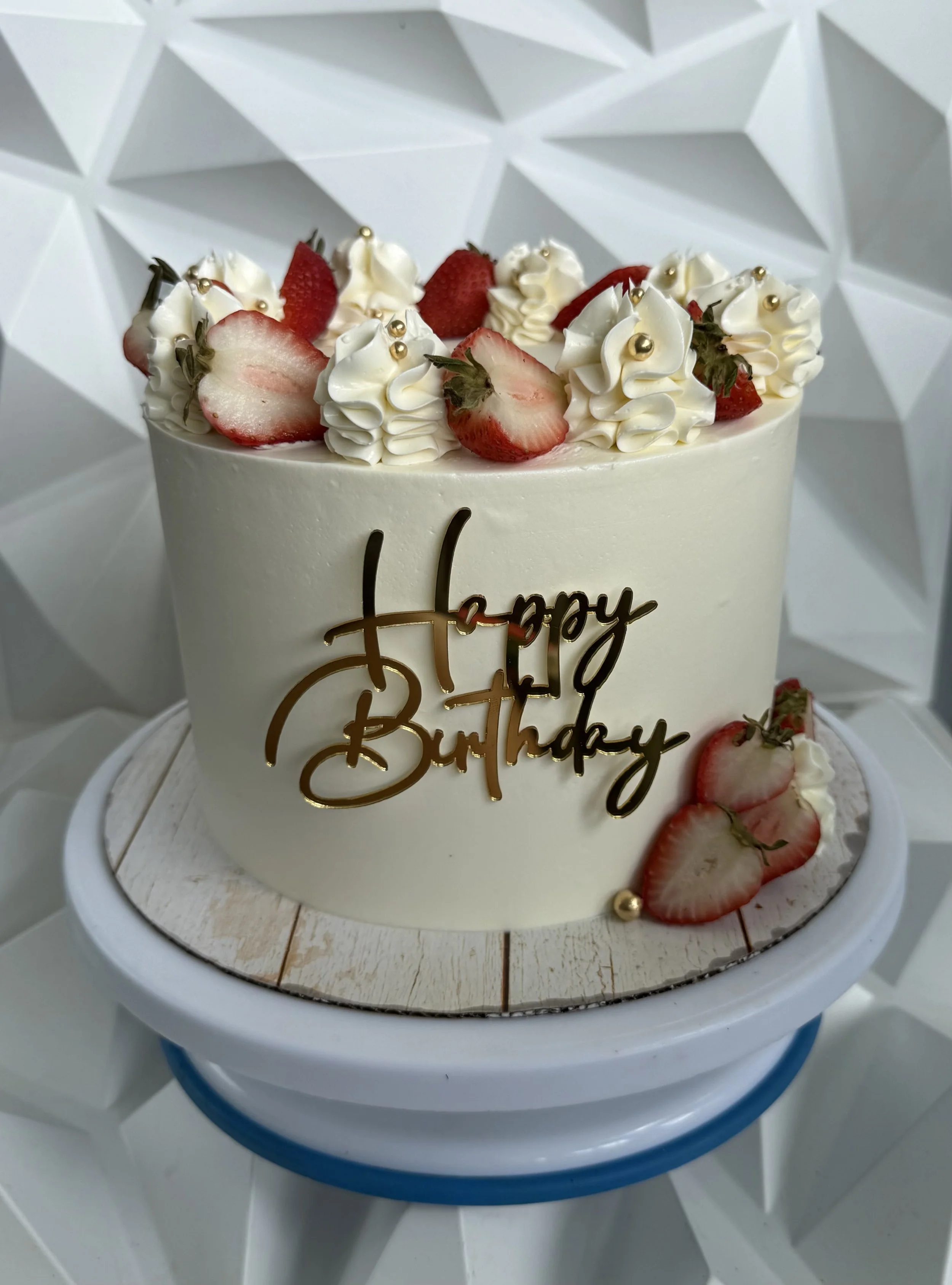 Pastel de cumpleaños con decoraciones de fresas, crema y perlas doradas, en una bandeja blanca sobre una mesa de madera.