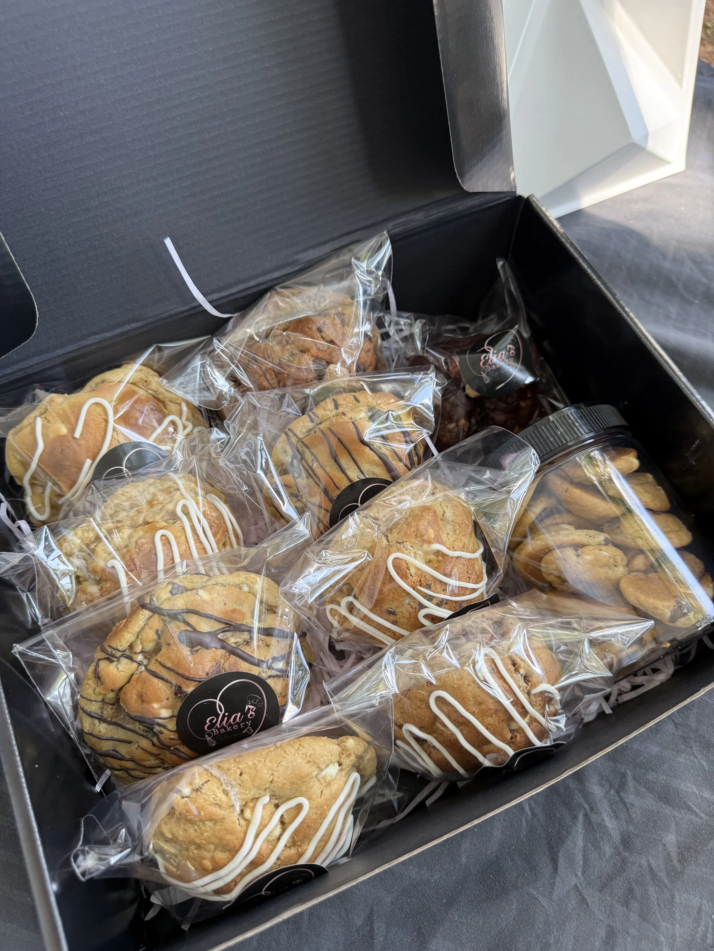 Caja con varias galletas empaquetadas en bolsas de plástico transparentes con etiquetas negras con letras y un logo, y algunos dulces y galletas sueltas en la caja.