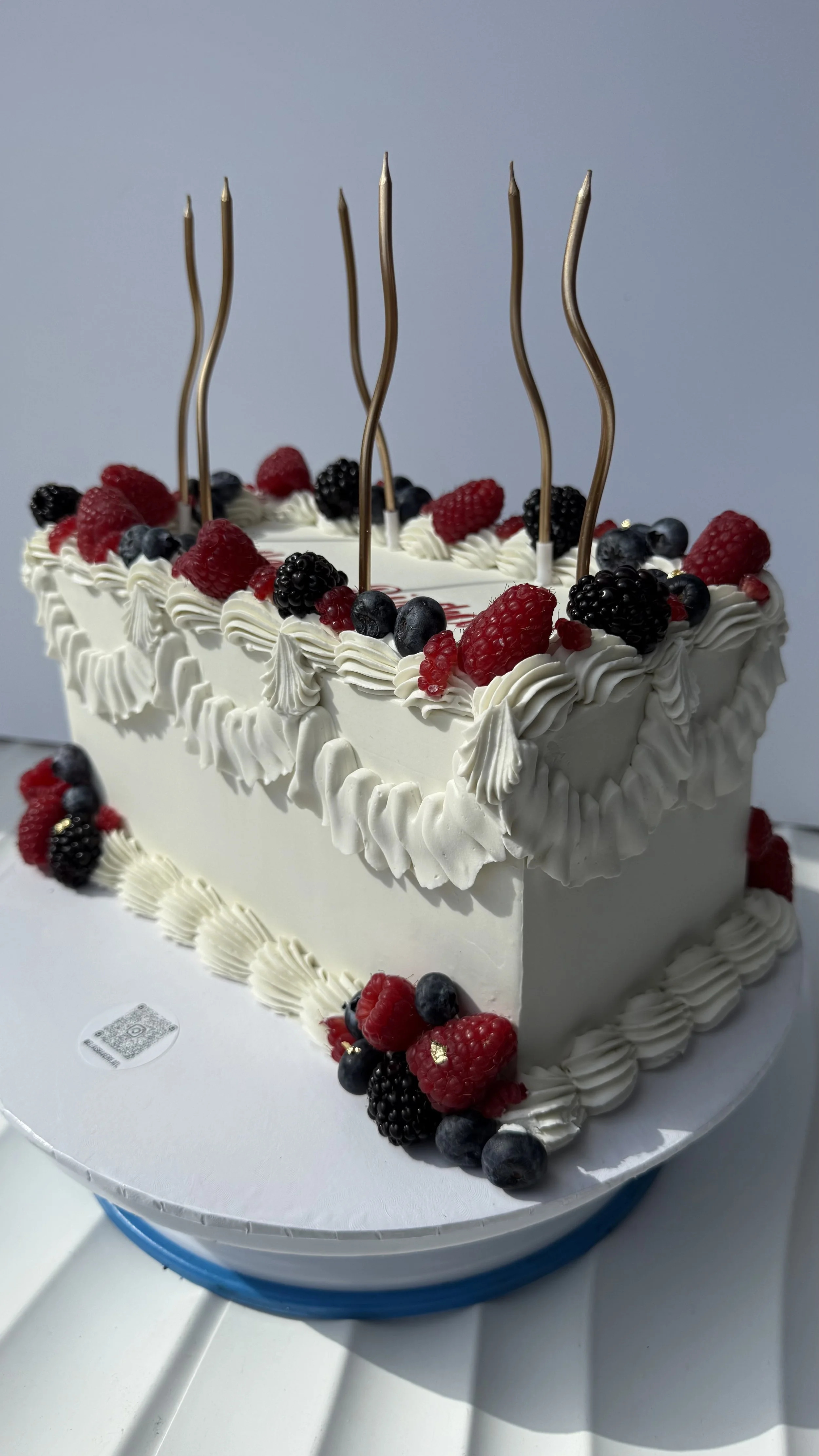 Pastel de cumpleaños rectangular decorado con crema, moras, frambuesas y arándanos, con cinco velas doradas en el centro y decoraciones con crema en los bordes.