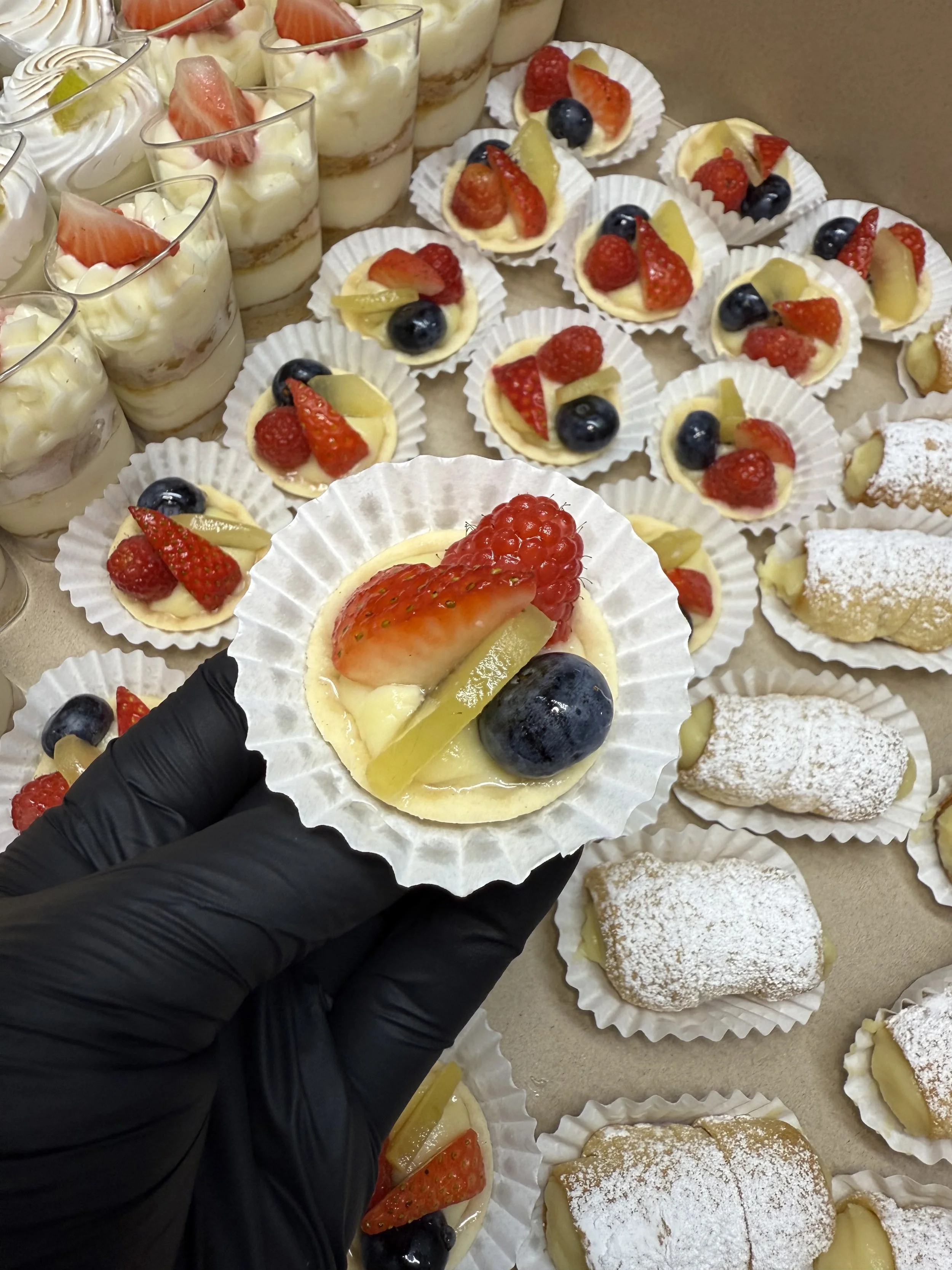 Variedad de postres con fresas, arándanos y otros frutos, algunos en copas de vidrio y otros en cápsulas de papel, en un mostrador.