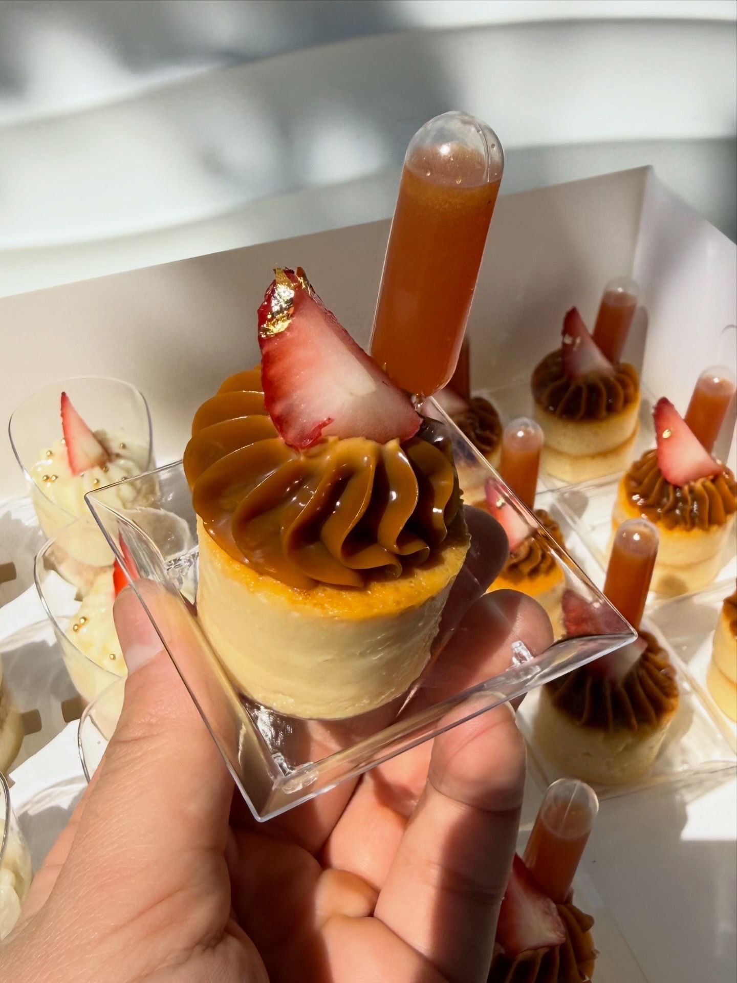 Postre en vaso con mousse de caramelo, trozos de fresa decorativos y un chupito de líquido ámbar, en un plato de presentación diferente con más postres similares.