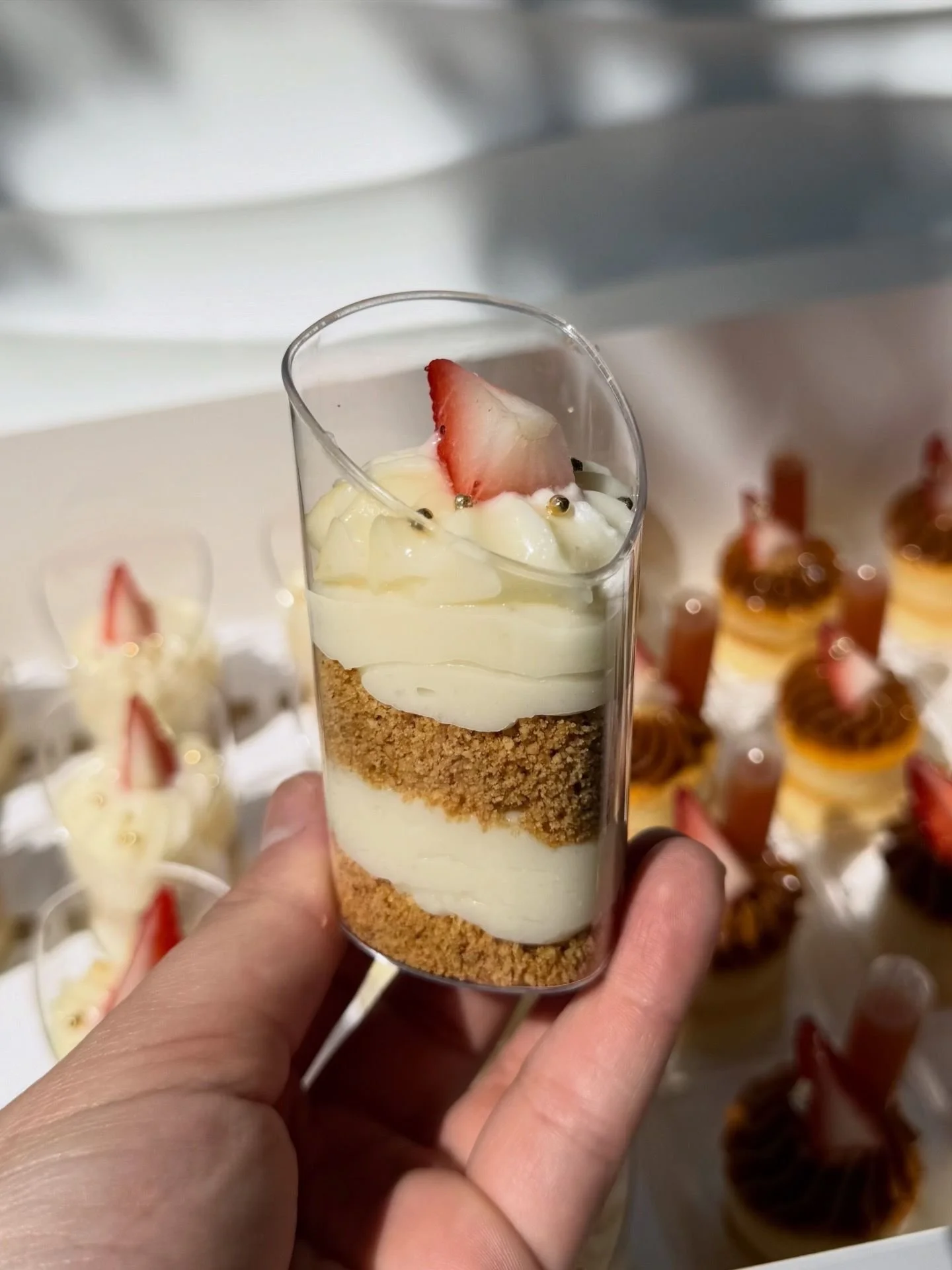 Vaso con postre en capas: base de galleta triturada, crema, parte superior decorada con crema, una fresita y pequeñas perlas doradas.