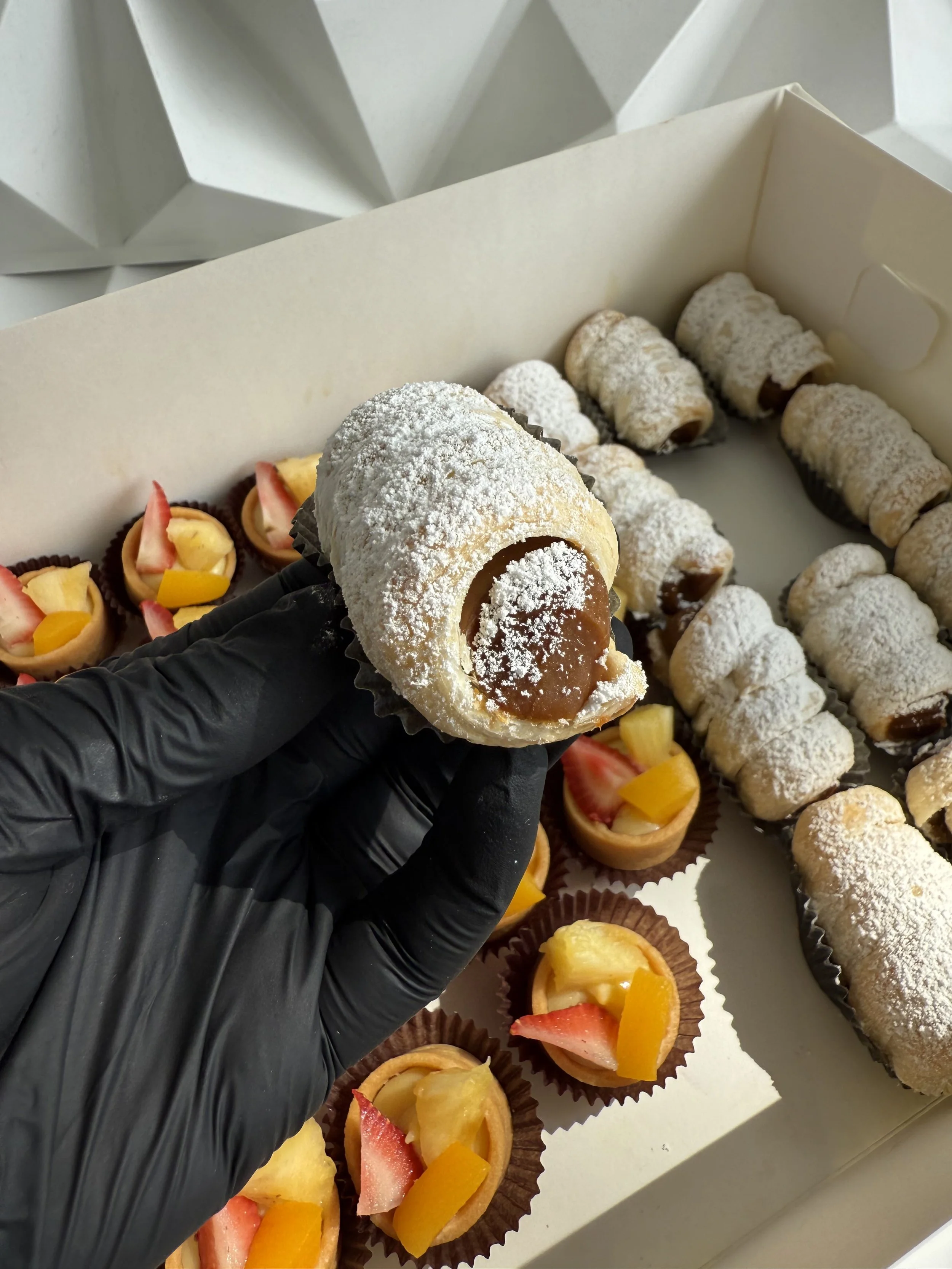 Persona con guantes negros sosteniendo un brazo de rollo de pastel relleno con chocolate y cubierto con azúcar en polvo, con variedad de pequeños pasteles y tartaletas con fruta en el fondo.