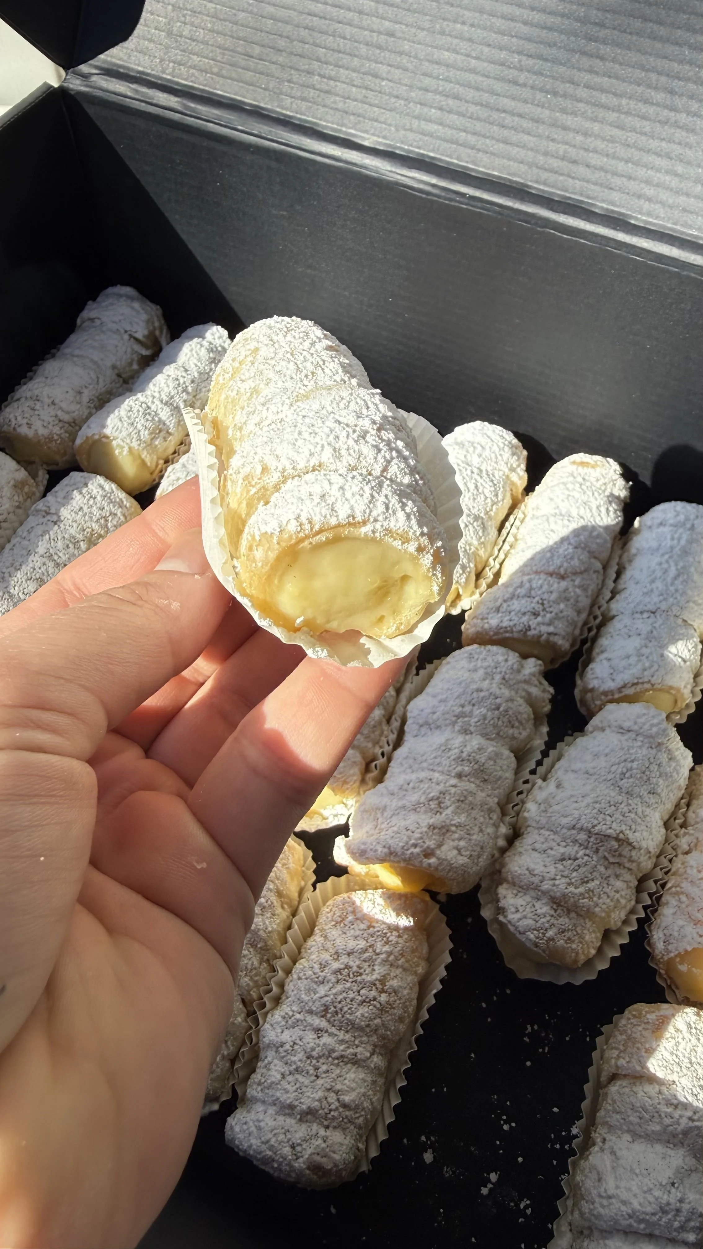 Varias rellenas en un recipiente negro cubiertas de polvo de azúcar, y una mano que sostiene una, mostrando su interior con crema o dulce