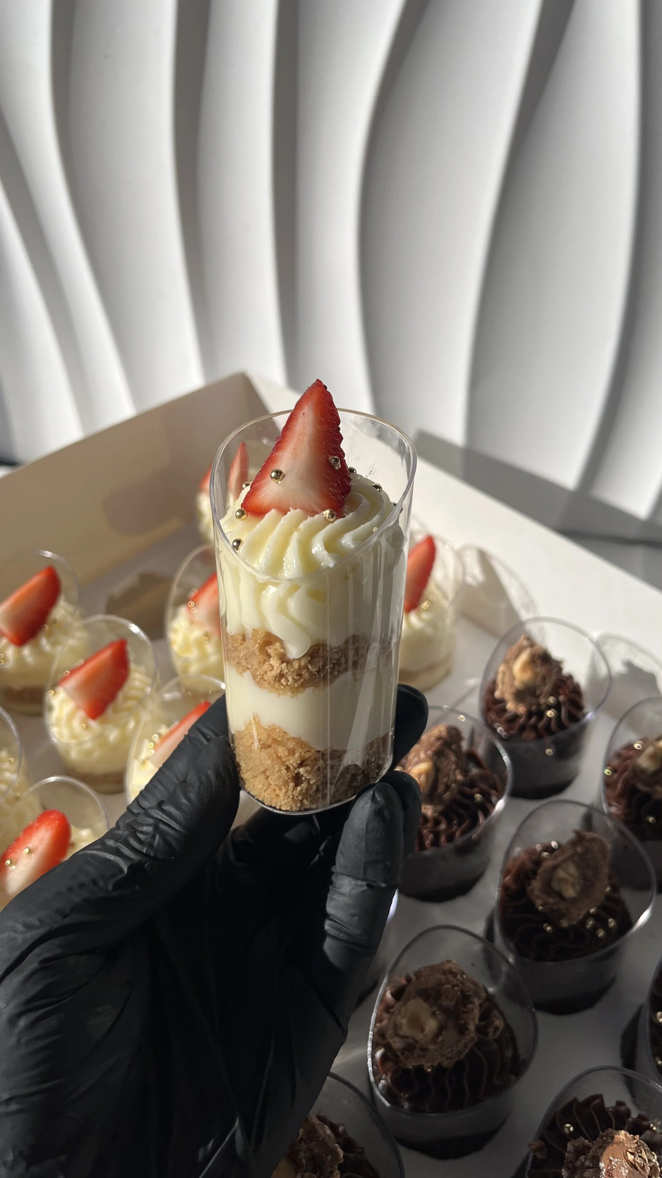 Vaso de postre con capas de crema, galletas trituradas y fruta de fresa en la parte superior, sostenido por una mano con guante negro, con otros postres en el fondo.