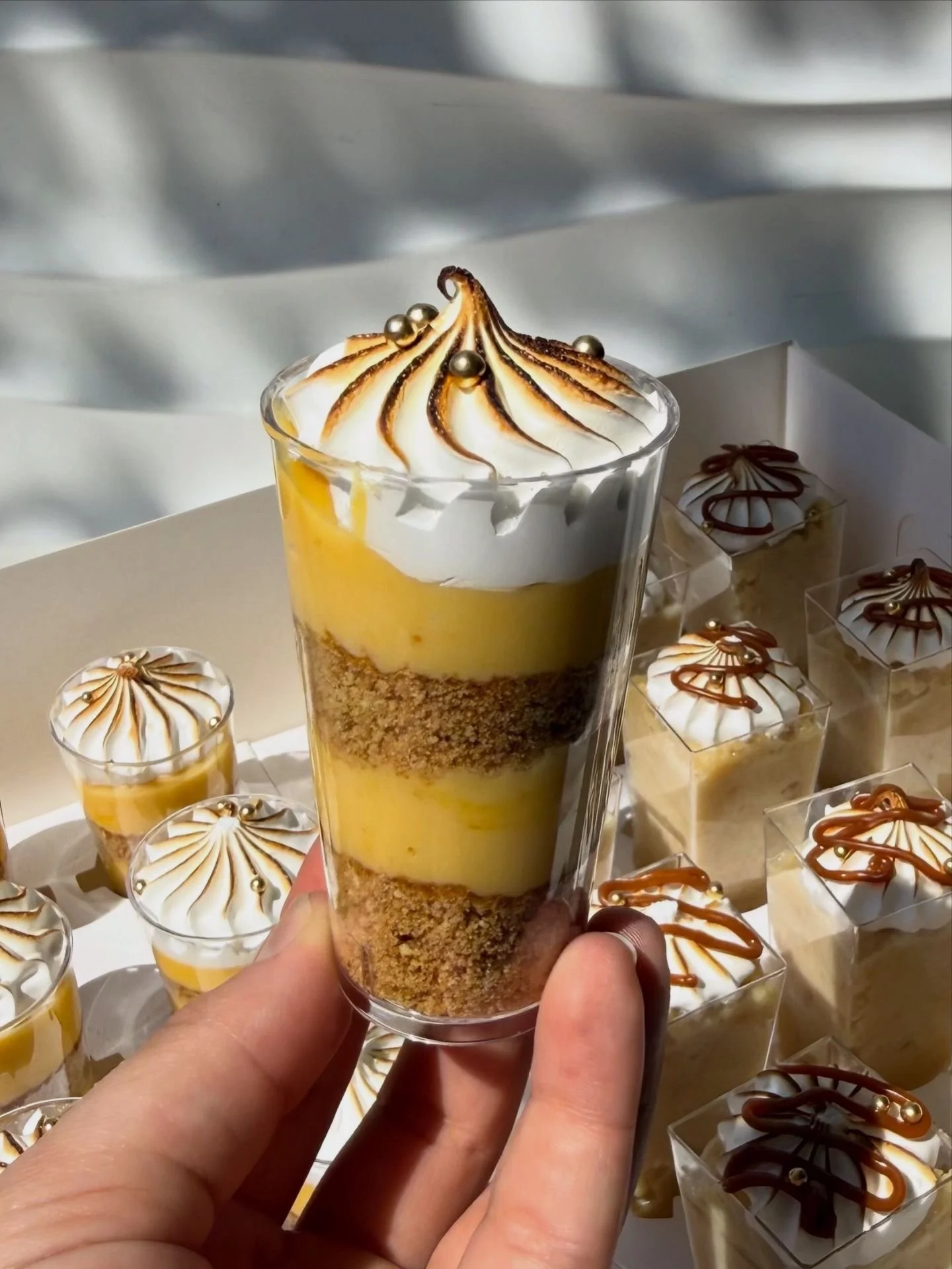 Vaso con postre de capas amarillas, de galleta y crema, con merengue tostado y adornos de bolitas doradas.