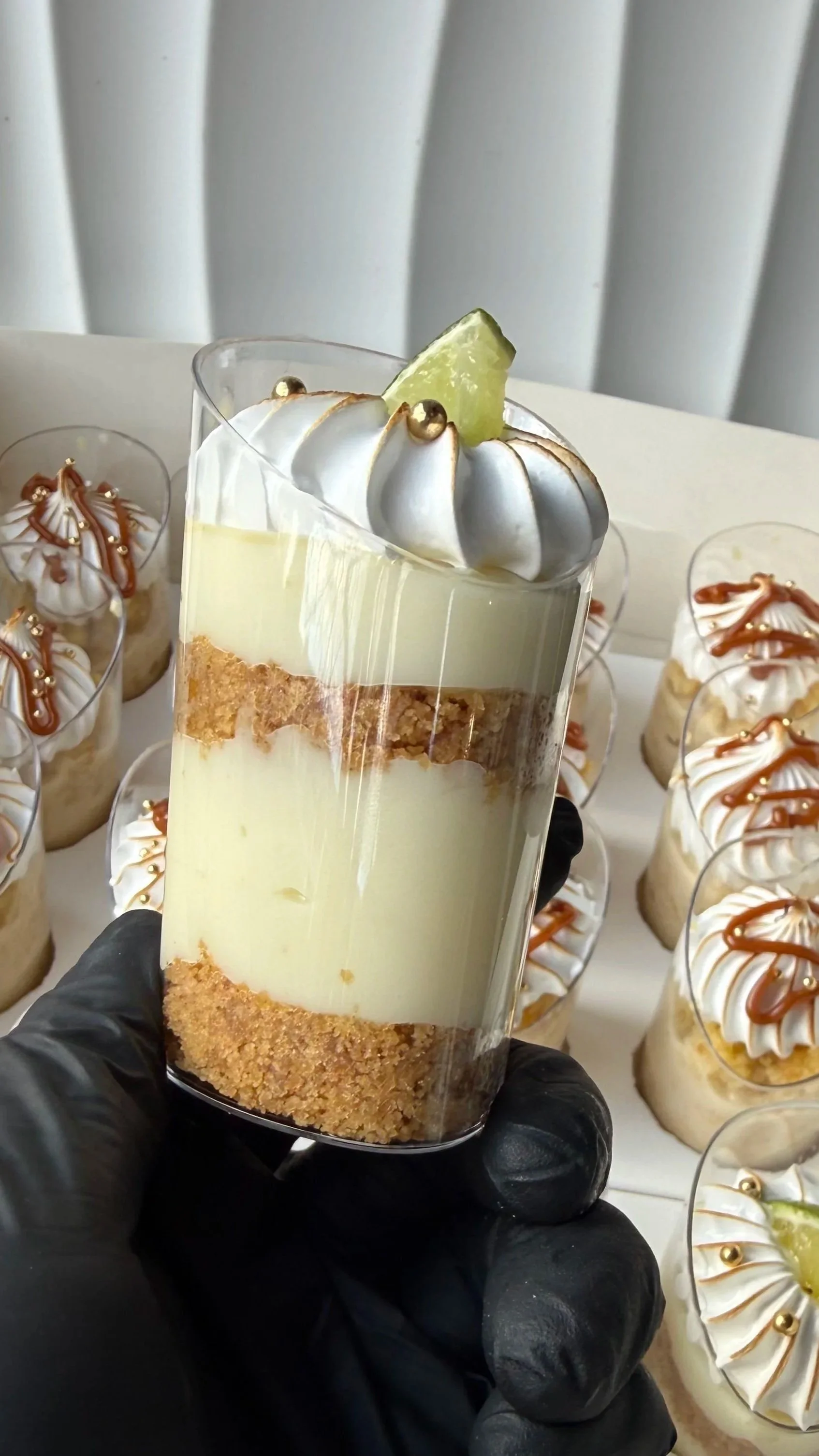 Vaso con postre en capas, decorado con una rodaja de limón y una bolita dorada, con otros postres similares al fondo.
