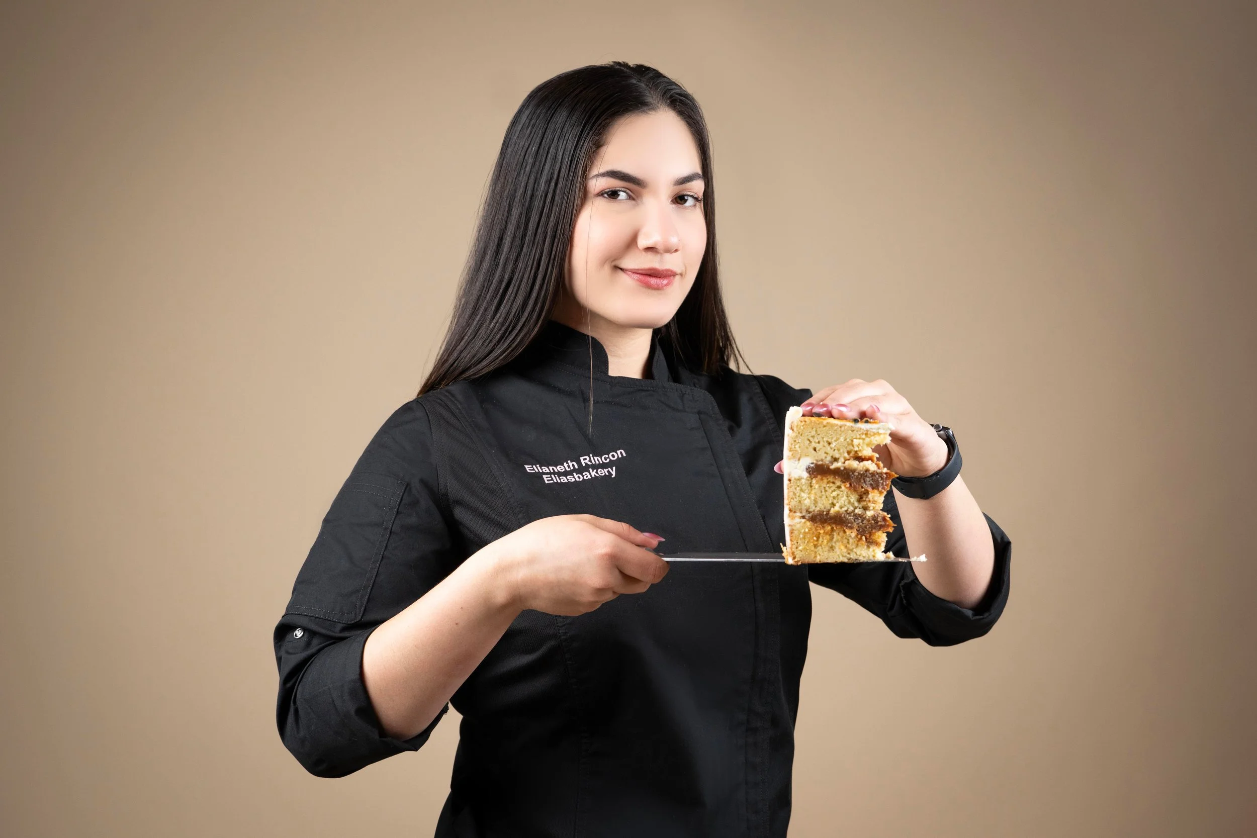 Mujer con bata de chef sosteniendo un trozo de pastel de varias capas y sabores, sonriendo a la cámara.