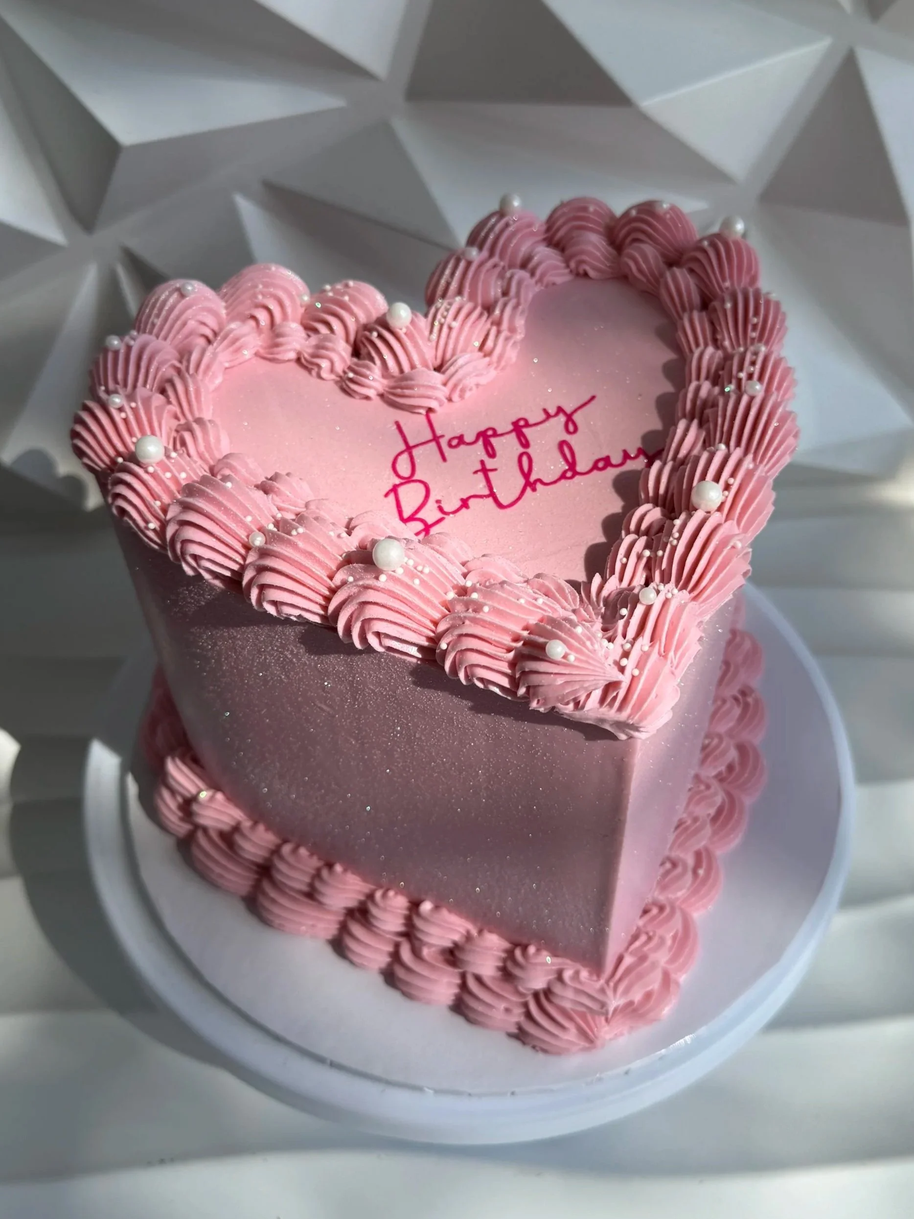 Pastel de cumpleaños en forma de corazón con decoraciones rosas y pequeñas perlas blancas, con la inscripción 'Happy Birthday' en el centro, en un plato blanco.