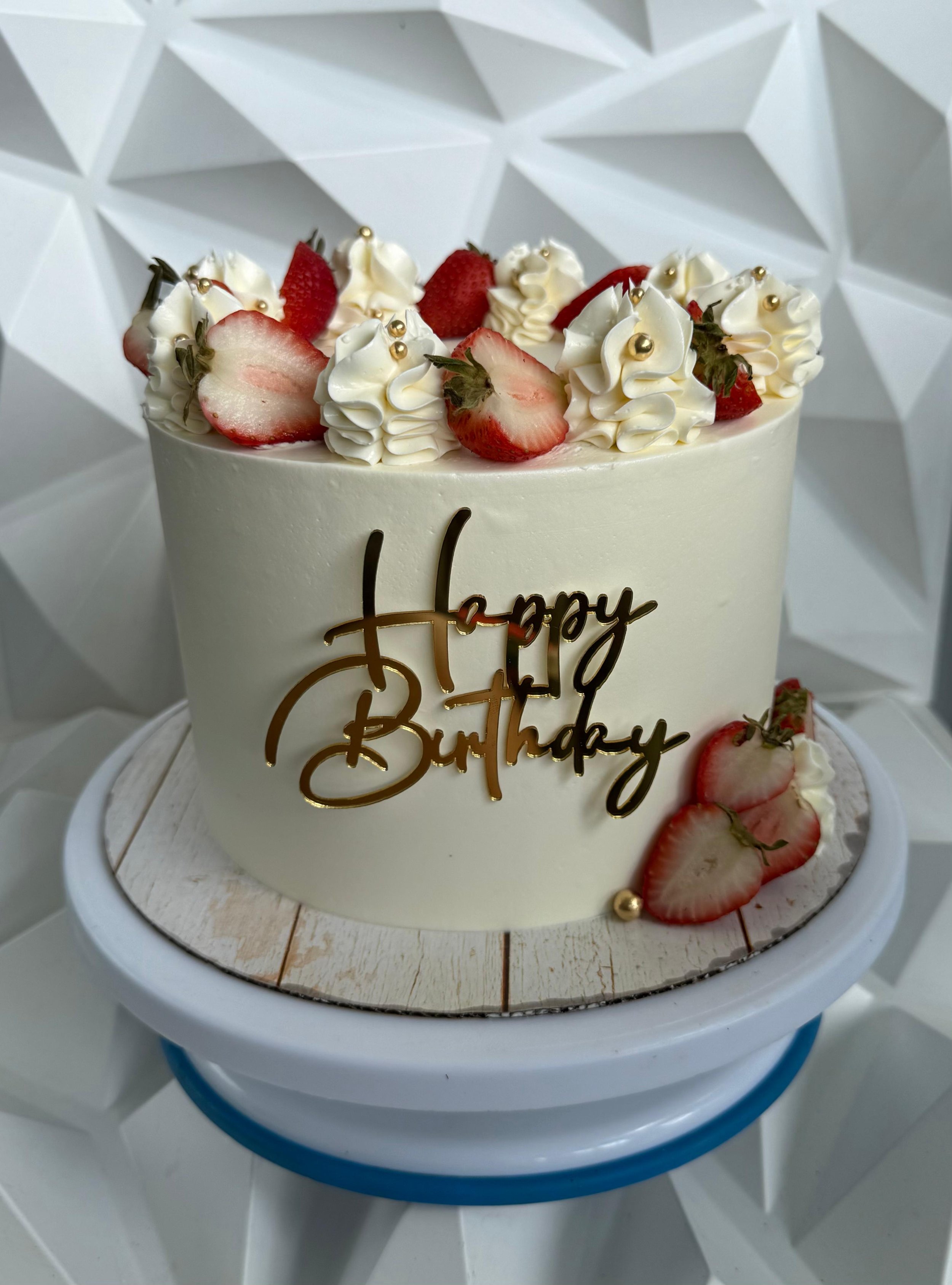 Pastel de cumpleaños decorado con crema, fresas y bolas doradas, con la palabra 'Happy' en letras doradas.