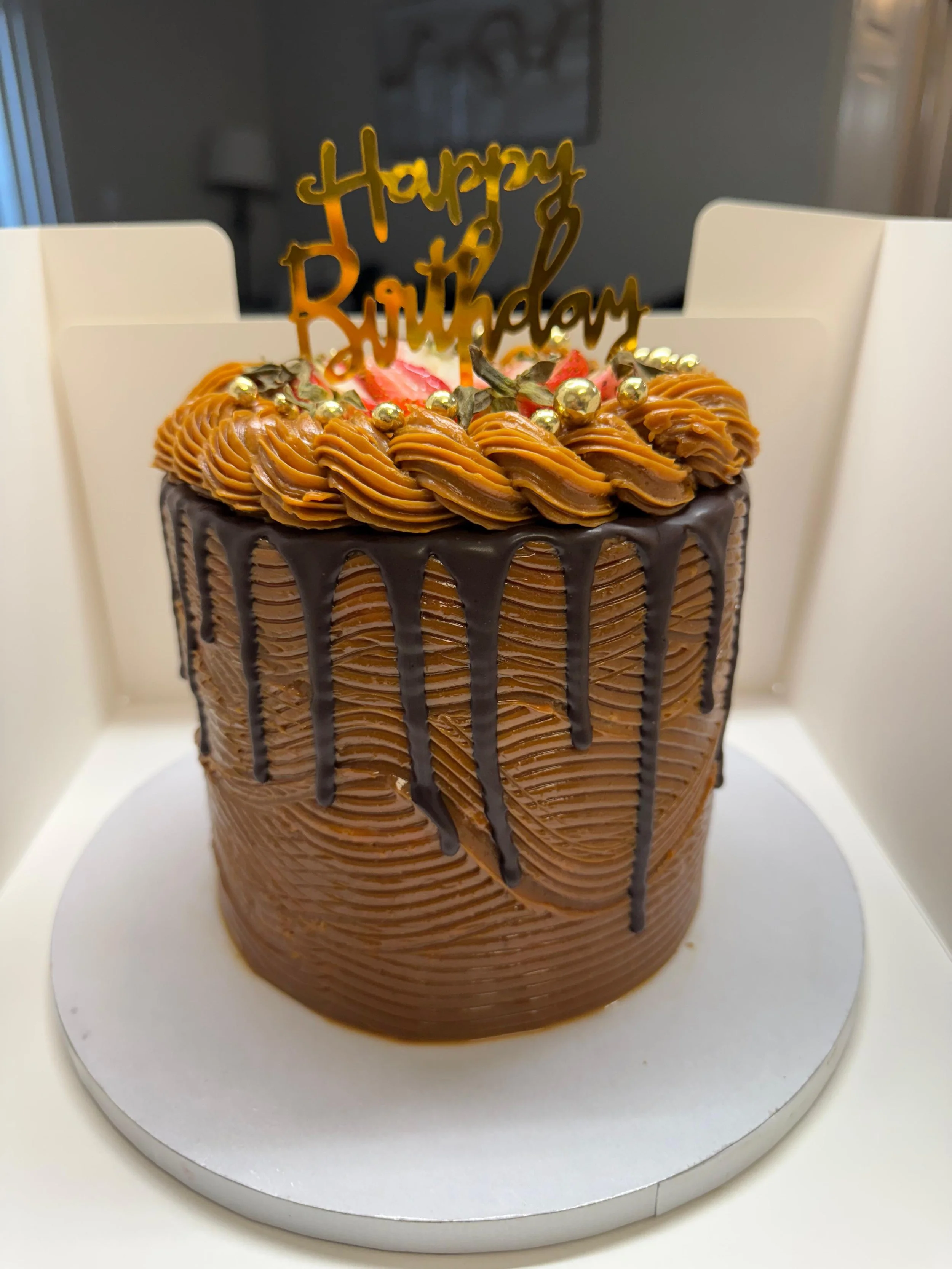Pastel de cumpleaños de chocolate decorado con cobertura de chocolate, crema de caramelo, chispas doradas y un topper que dice 'Feliz Cumpleaños' en estilo dorado, sobre una base blanca
