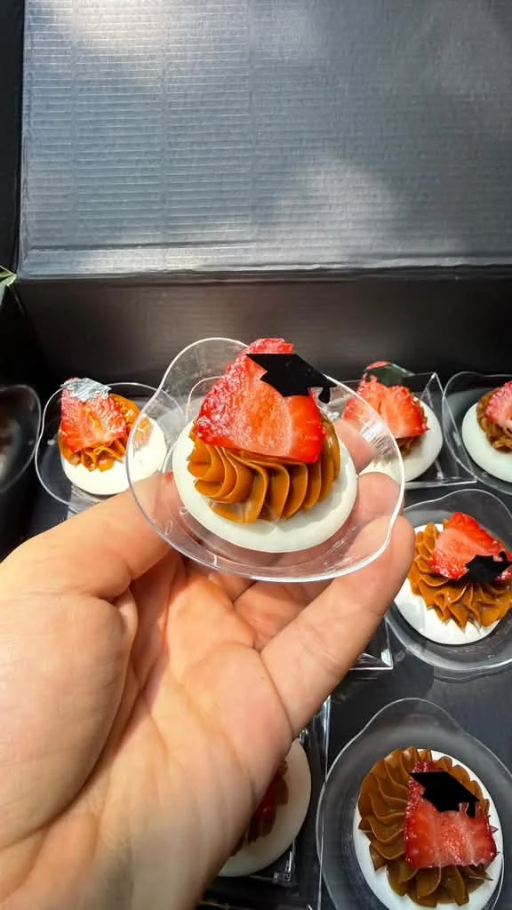 Postre envasado en una copa plástica, con crema, chocolate, fresa y decoraciones negras.
