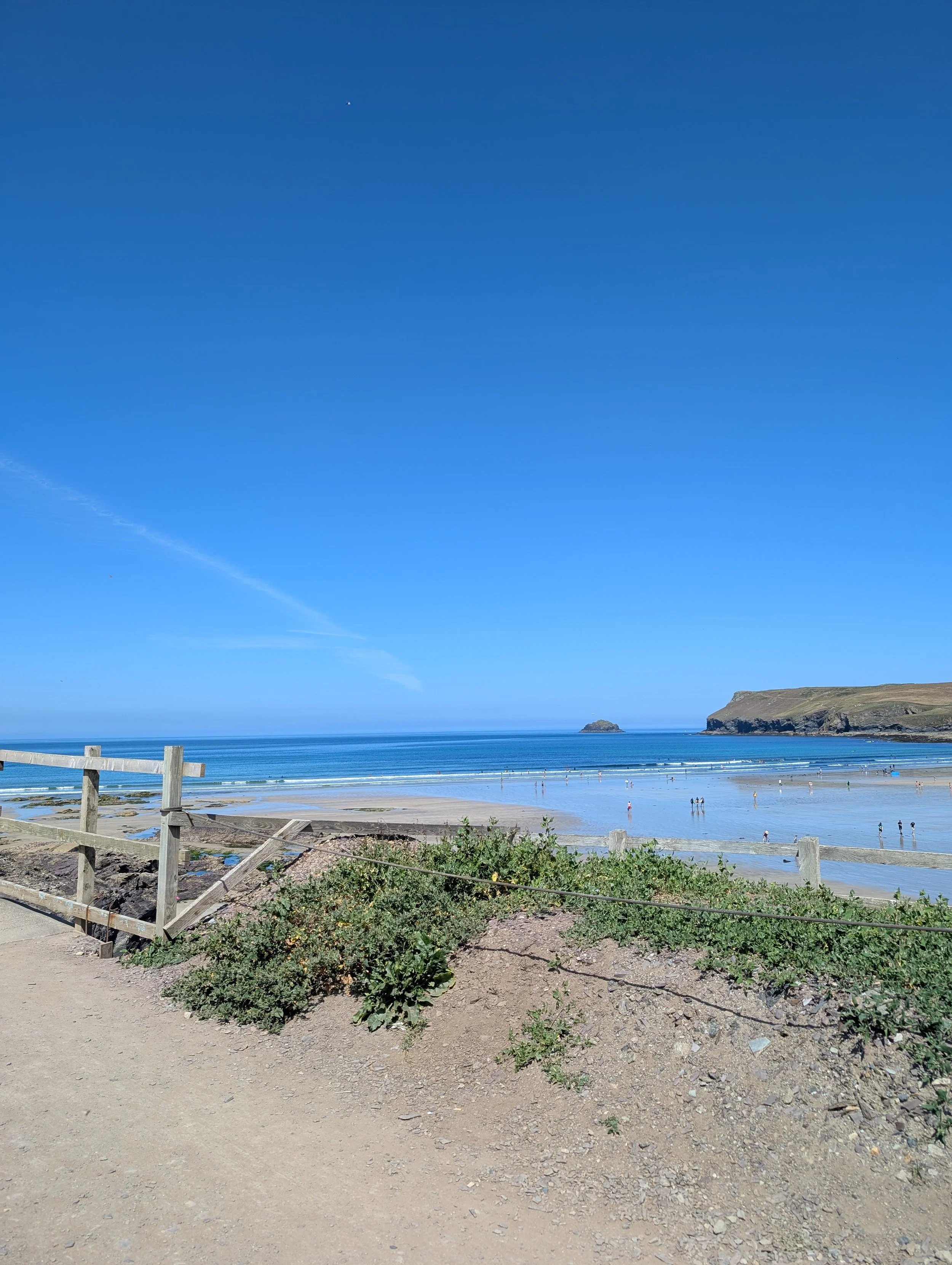 Sunny view over Polzeath Beach, Cornwall, with blue skies, calm sea and stepper point on the horizon.
