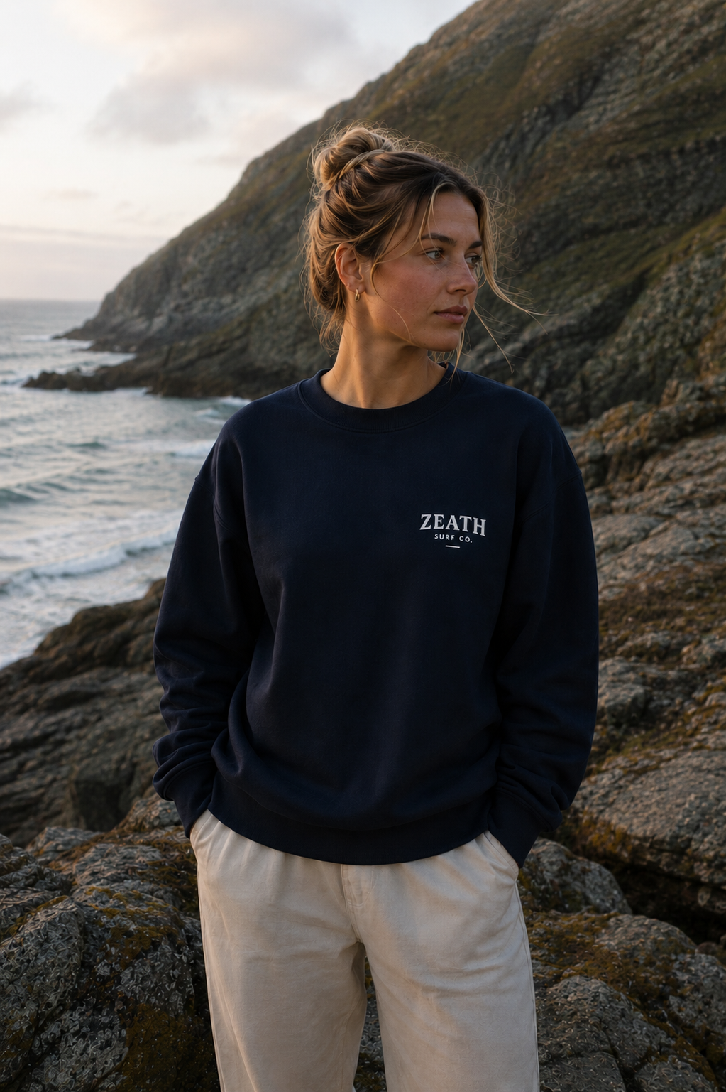 A woman standing on rocky cliffs near the ocean at sunset, wearing a navy sweatshirt with 'ZEATH SURF CO.' logo embroidered in white.