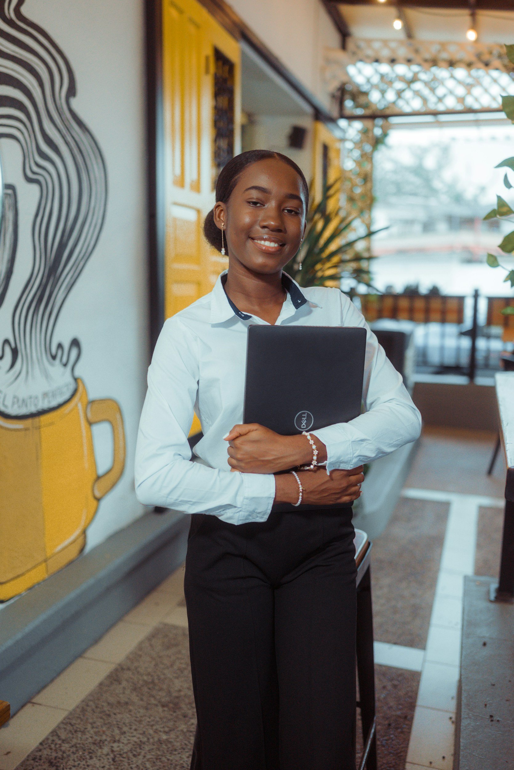 A smiling woman wearing a white blouse with a dark collar, holding a closed laptop, standing inside a cafe or restaurant near a yellow wall with beer-related artwork.