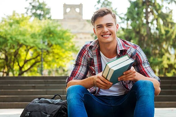 A young man sitting on outdoor steps, holding books, smiling, with a backpack beside him, trees and a building in the background.