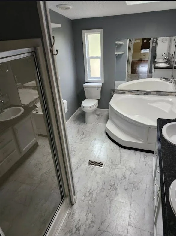 Bathroom with gray walls, white toilet, window, corner bathtub with a curved edge, double sinks, and a glass-enclosed shower. Marble-style floor tiles.