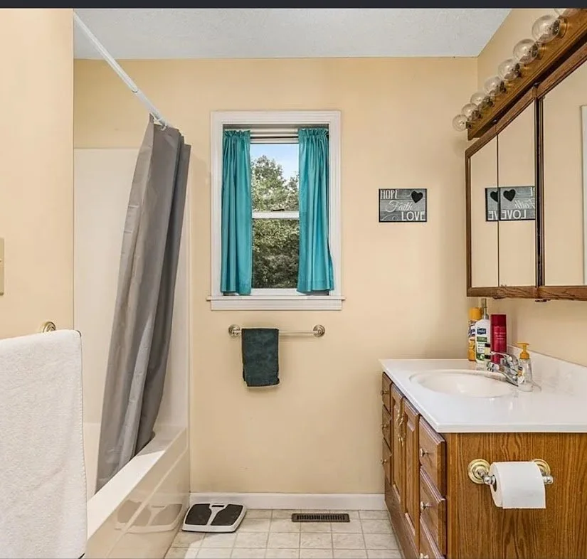 Bathroom with beige walls, a small window with blue curtains, a wooden vanity with a mirror, and a bathtub with gray shower curtain. There are toiletries on the sink, a towel on a rack, a scale, and a small decorative sign on the wall.