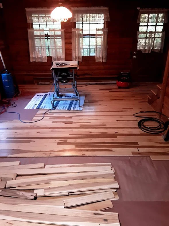 Room with wooden floors and walls, two windows with lace curtains, table saw on a small stand, tools, and wooden planks on the floor indicating renovation work.