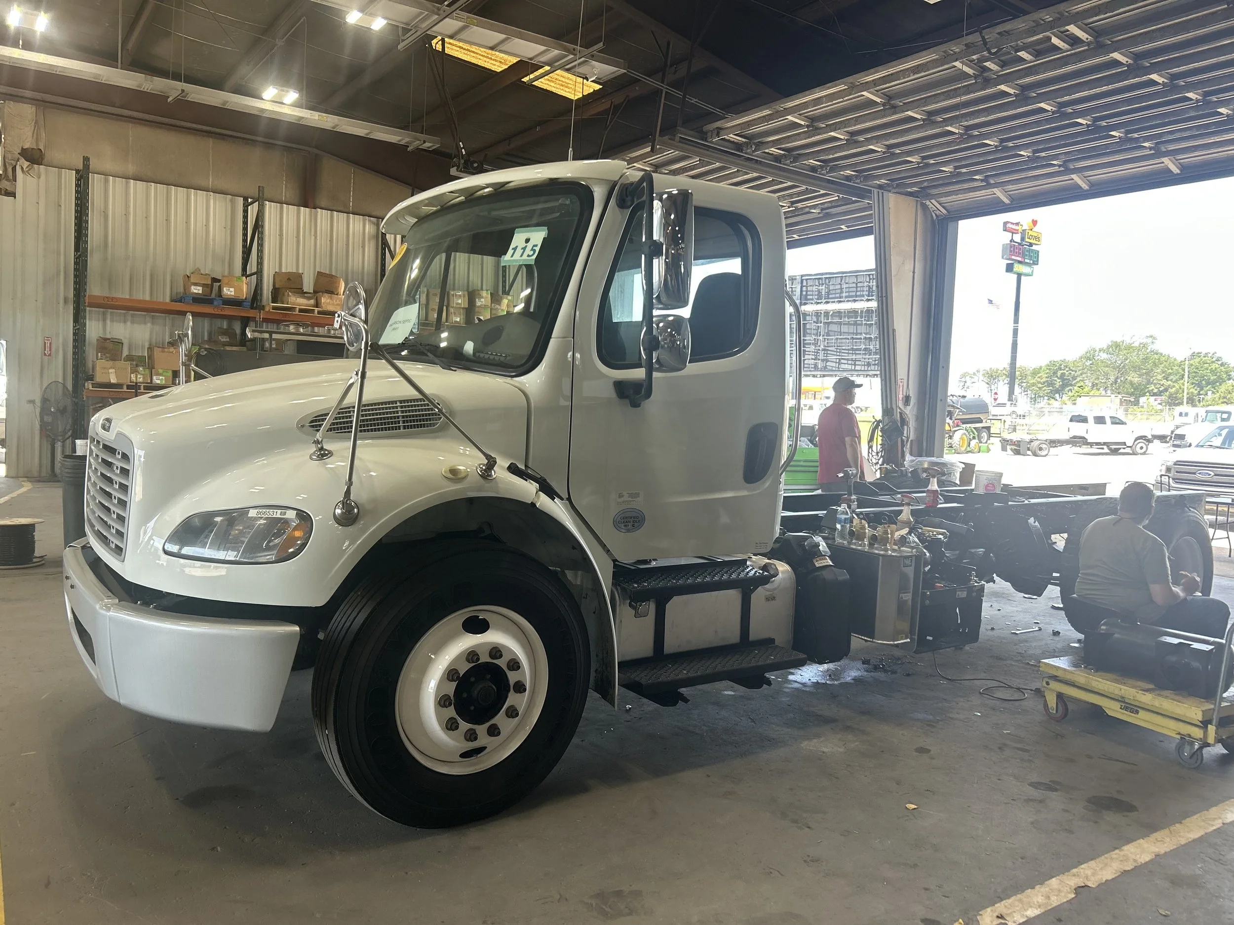 Inside a garage or auto shop with a large white semi-truck cab. The truck is in the process of being serviced or assembled, with parts visible, including a new front wheel and various tools and equipment around. There are shelves with boxes and a per