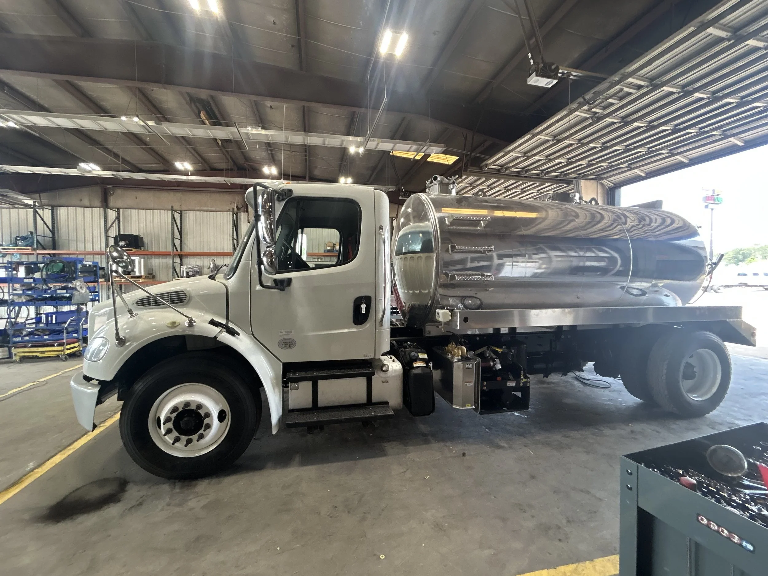 A silver tanker truck inside a warehouse with a metal roof and shelves in the background.