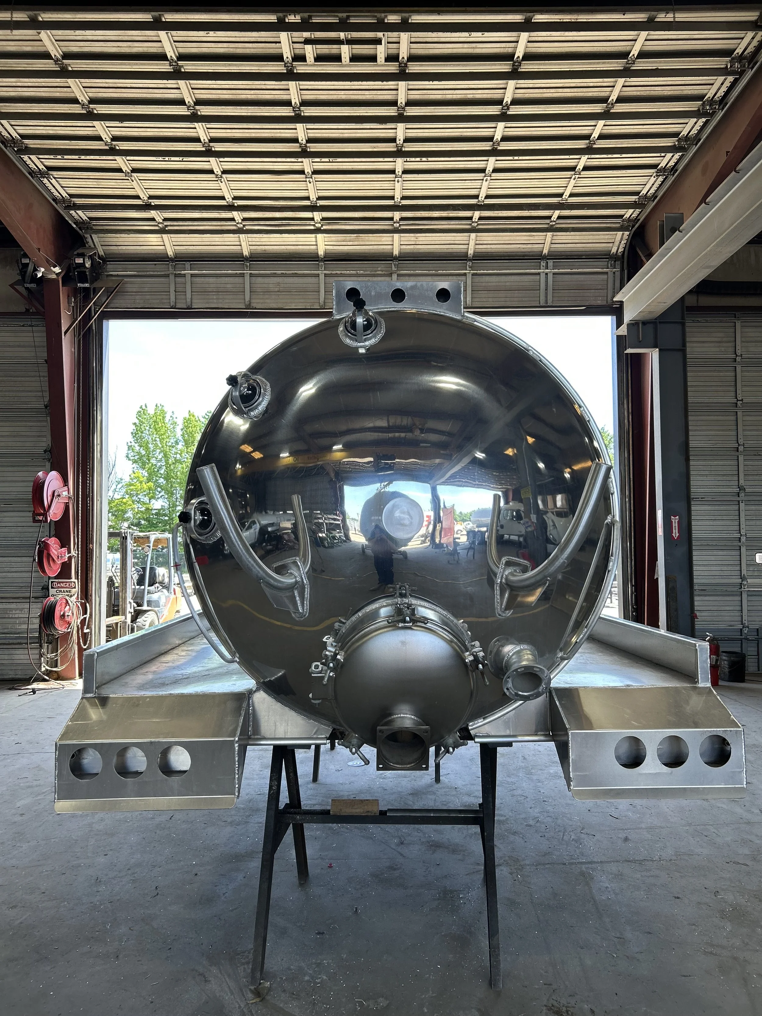 Front view of a large stainless steel industrial tank on a work table inside a warehouse, with tools and equipment around.