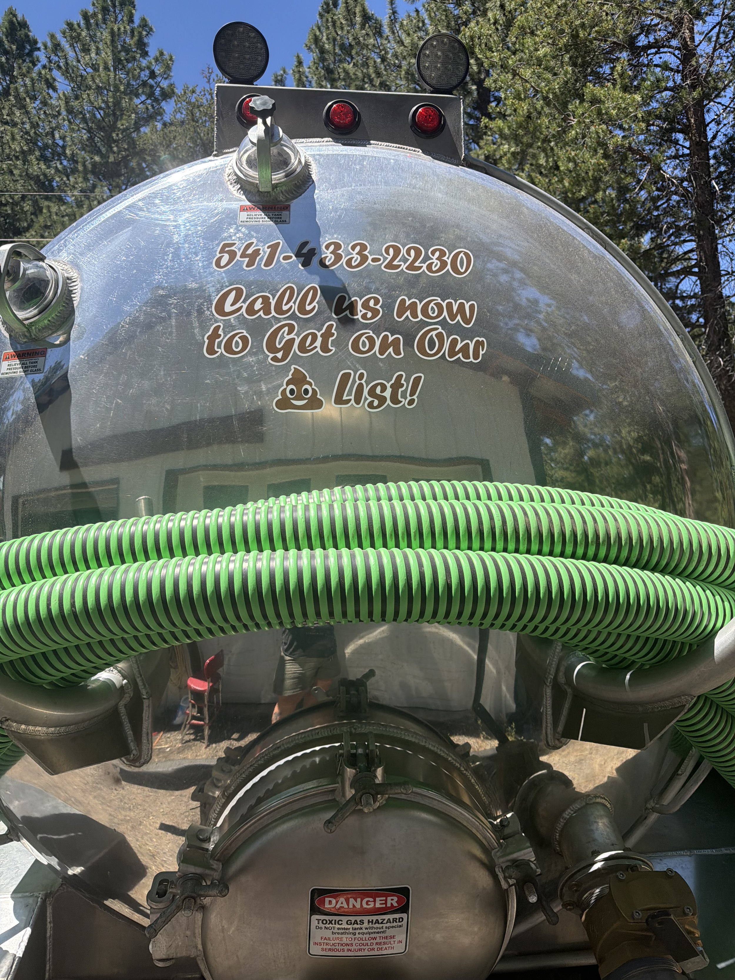 Close-up of a silver tank with a green and black coiled hose wrapped around it, labeled with a warning sign about toxic gas hazard, with trees and blue sky in the background.
