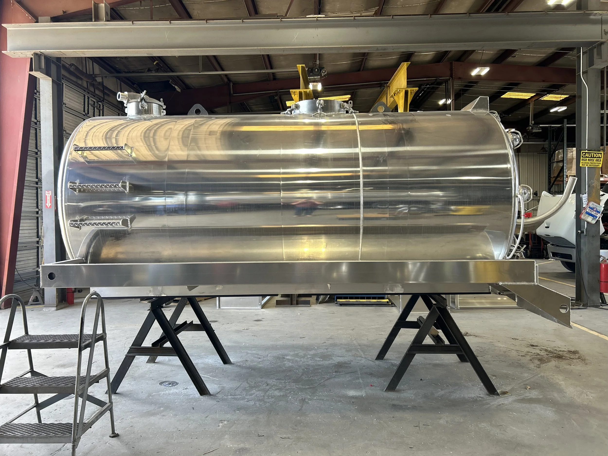 Large stainless steel industrial tank or vessel inside a warehouse or factory with metal framework and equipment around it.