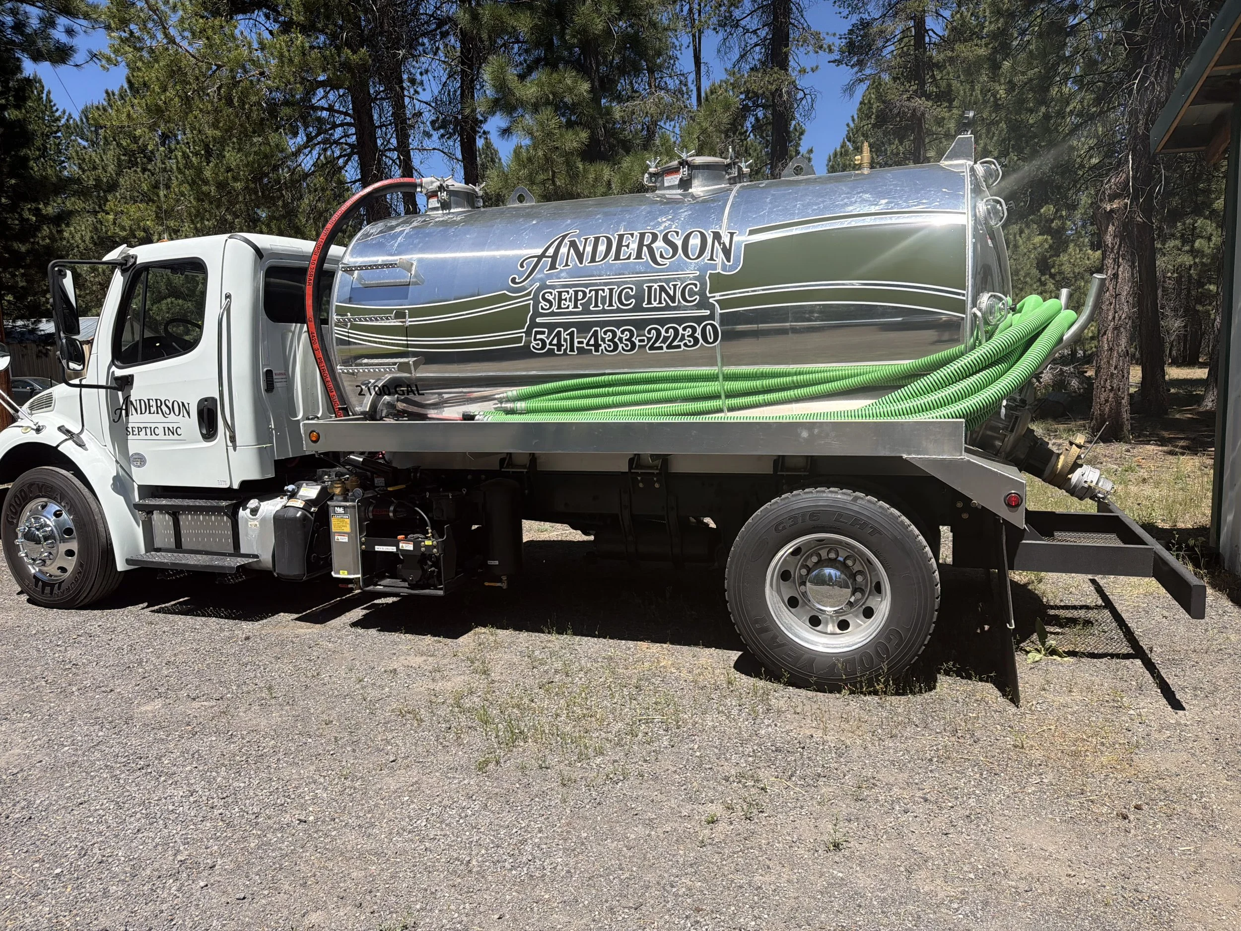 A white truck with a large metal septic tank on its back, branded 'Anderson Septic Inc' with a phone number. The tank has several green hoses and safety labels, and the scene is outdoors with trees and blue sky in the background.