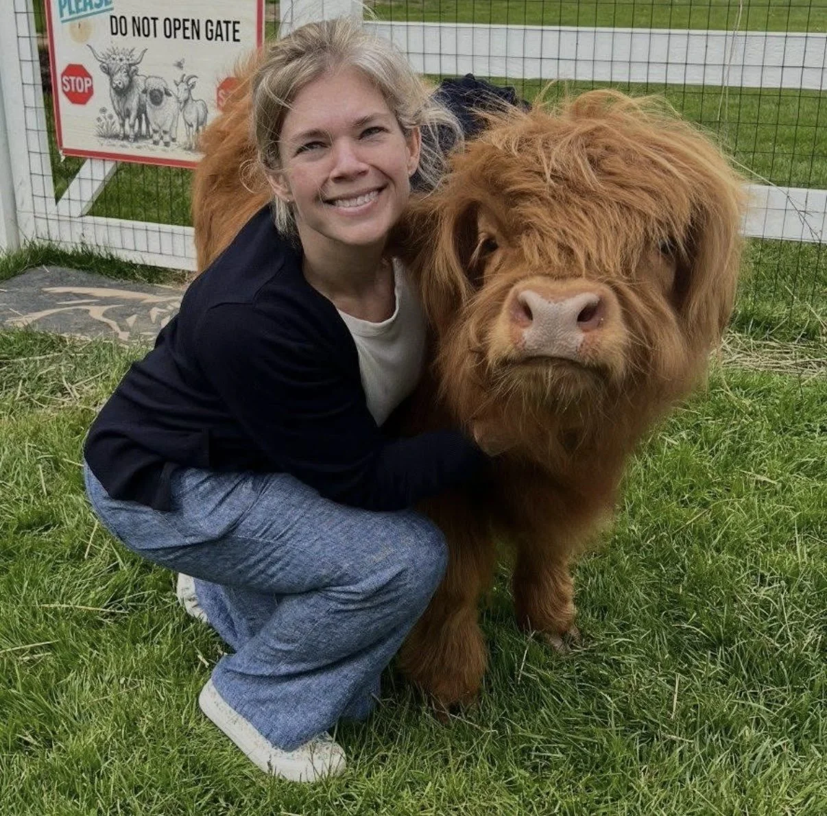 A woman crouching on green grass, smiling, next to a large brown Highland cow with long shaggy hair, in an outdoor fenced area.