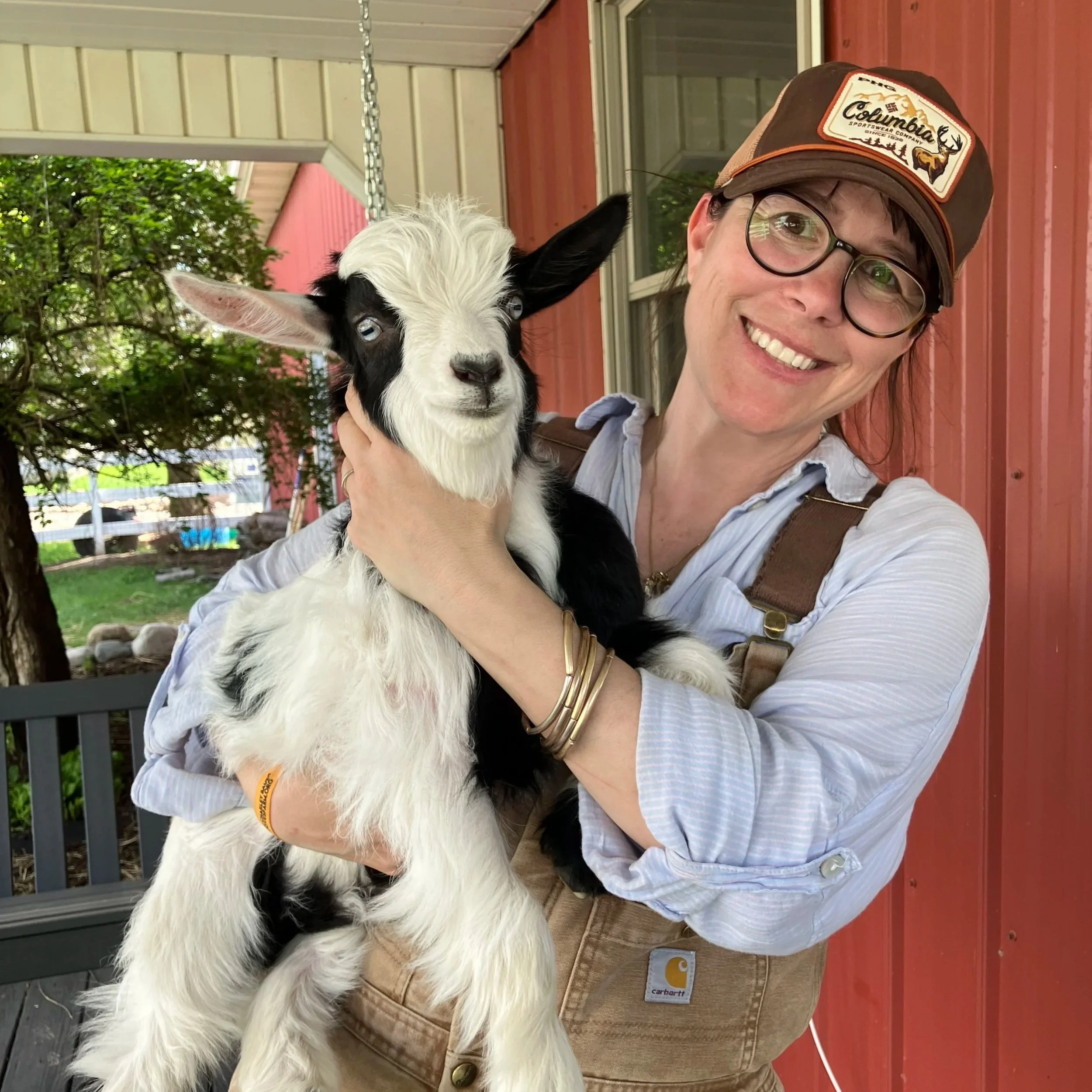 A woman smiling and wearing glasses, a cap, and a light blue shirt, holding a black and white baby goat, outdoors near a house with red siding and a window, trees in the background.