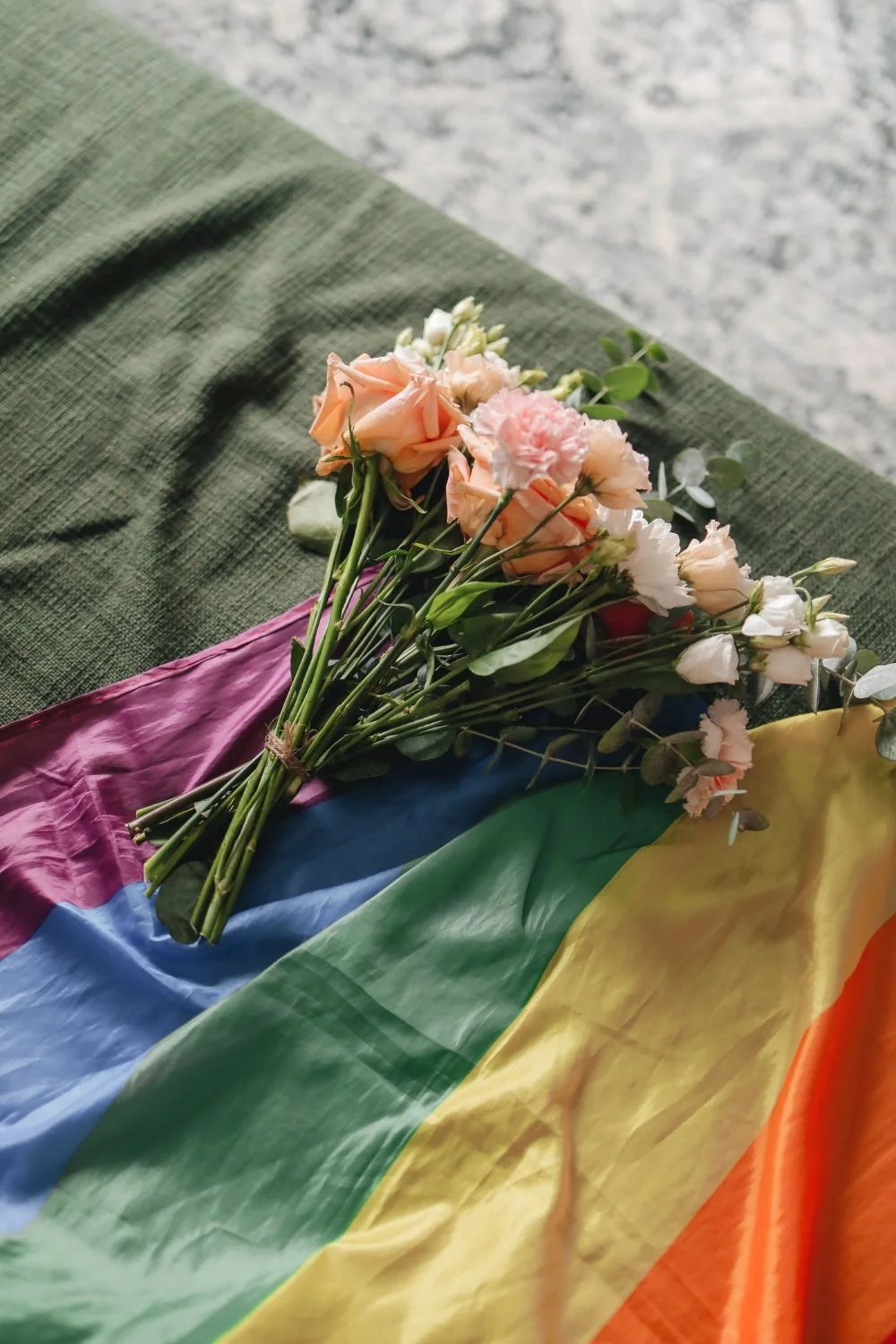 A bouquet of pink, white, and peach flowers resting on a multicolored rainbow flag on a green textured fabric surface.