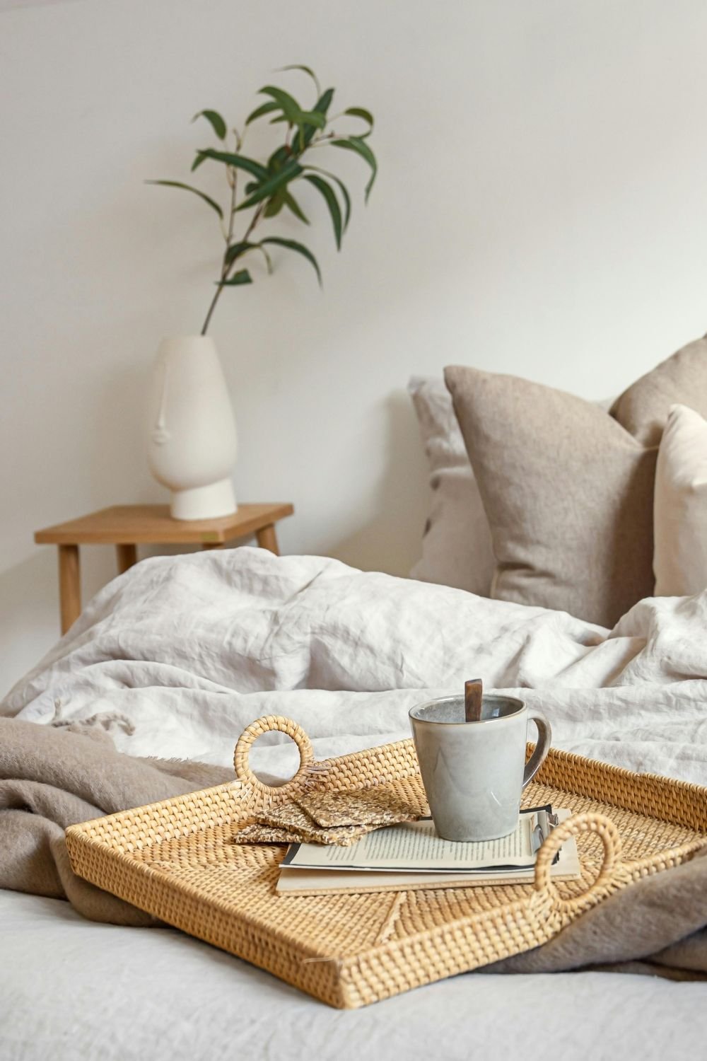 A cozy bedroom with a bed having beige and cream pillows, a wooden nightstand with a white vase and green plant, a woven tray with a mug, snacks, and books placed on the bed.