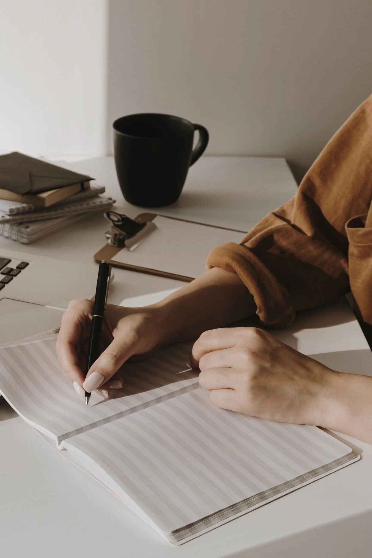 Person writing in a notebook at a white desk with a black coffee mug and stacks of books beside it.
