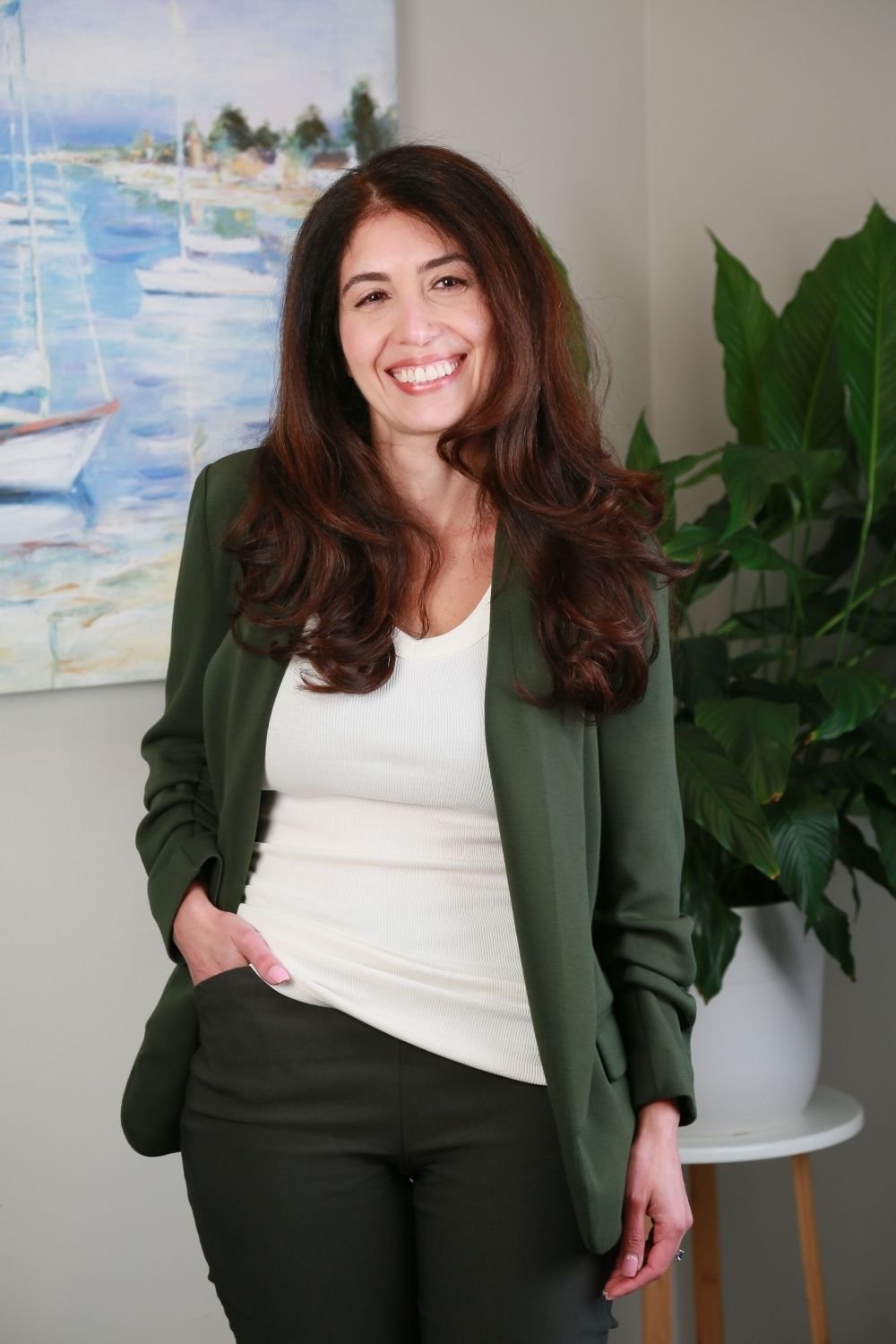 A woman with long, wavy brown hair smiling and standing indoors in front of a painting of sailboats and a large green plant.