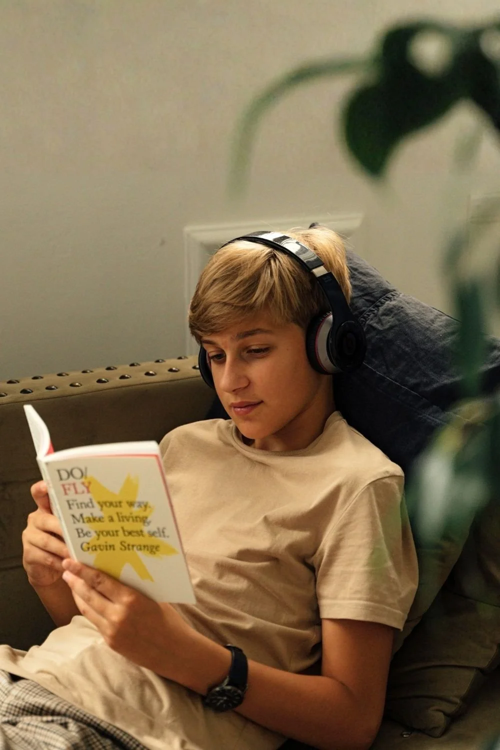 A teenage boy sitting on a couch, wearing headphones, reading a book about finding your way and being your best self, with a plant partially visible in the foreground.