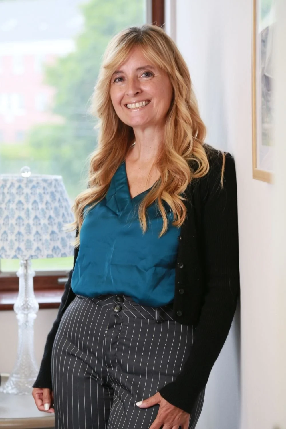 A woman with long, wavy strawberry blonde hair, smiling and standing near a window with a view of trees and buildings outside, wearing a teal blouse, black cardigan, and striped pants.