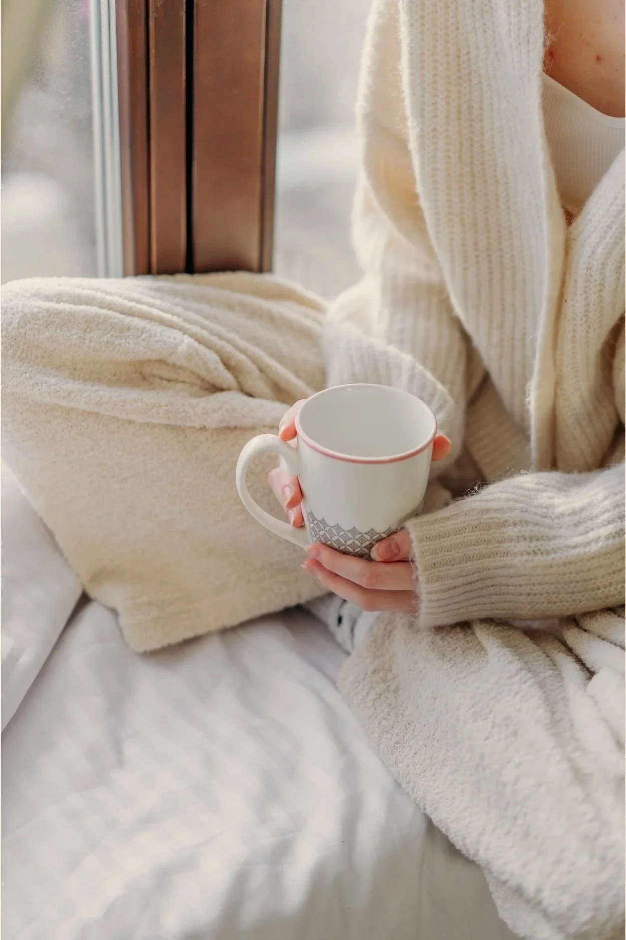 Person sitting on a bed wearing a cream-colored sweater, holding a white mug with a decorative pattern, with a beige blanket draped over their lap and a window in the background.