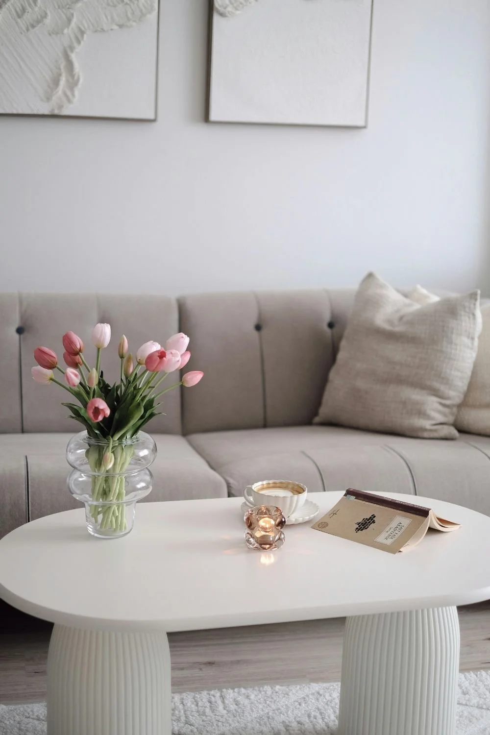 Living room with a beige sofa, beige pillow, white coffee table with a vase of pink tulips, a cup of coffee, a small glass candle holder, and an open book.