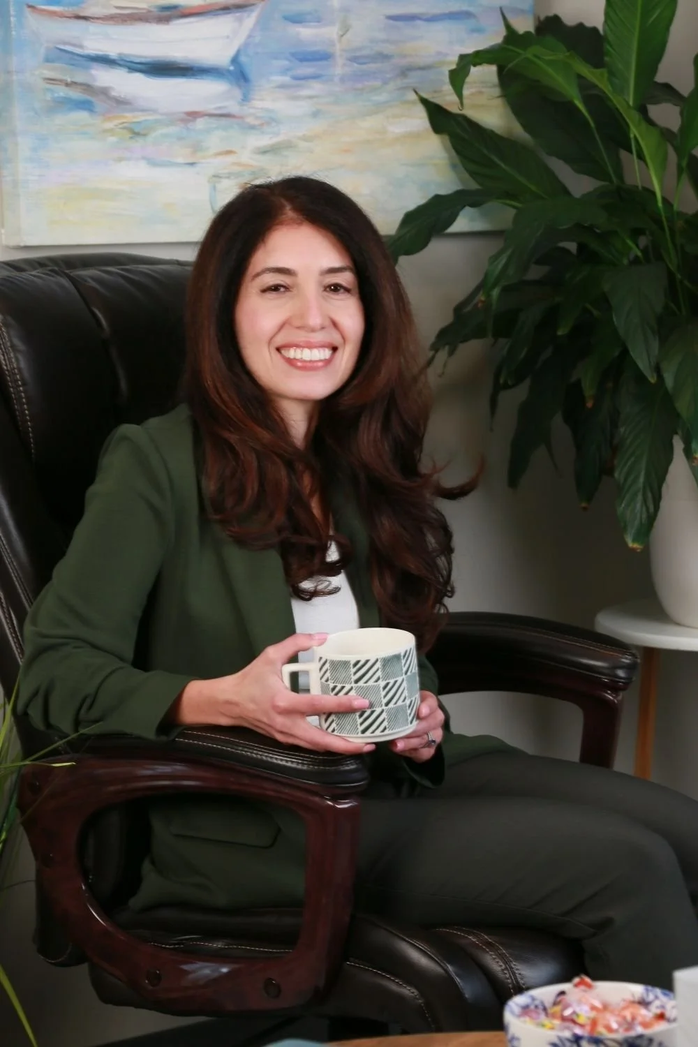 A woman with long dark hair smiling while sitting in a leather chair, holding a patterned mug, with a large green plant and a painting of a boat in the background.