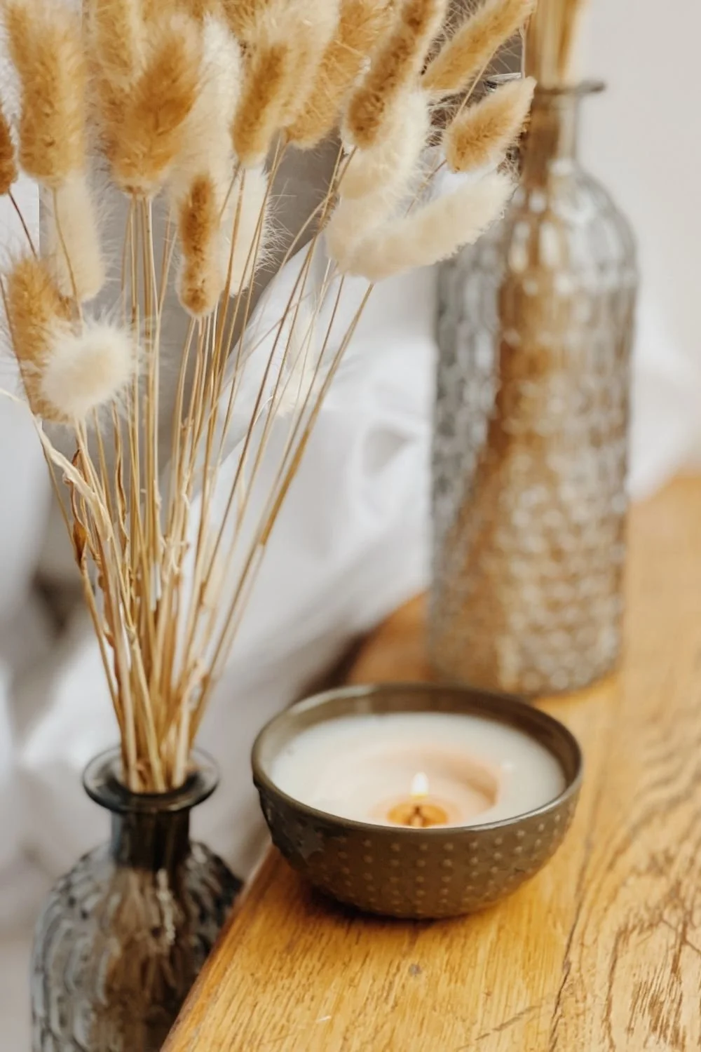 Dried beige and white pampas grass in a black glass vase, a black dotted candle holder with a lit candle, and a tall textured glass bottle on a wooden surface.