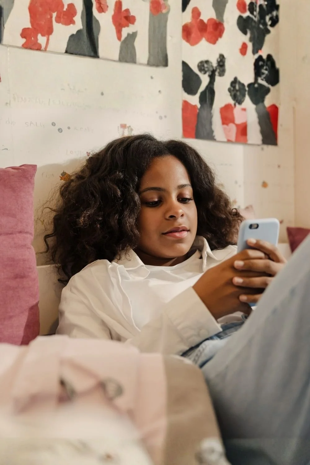 A young girl with curly hair lying on a bed, looking at her phone with a focused expression, in a room with artwork on the wall behind her.