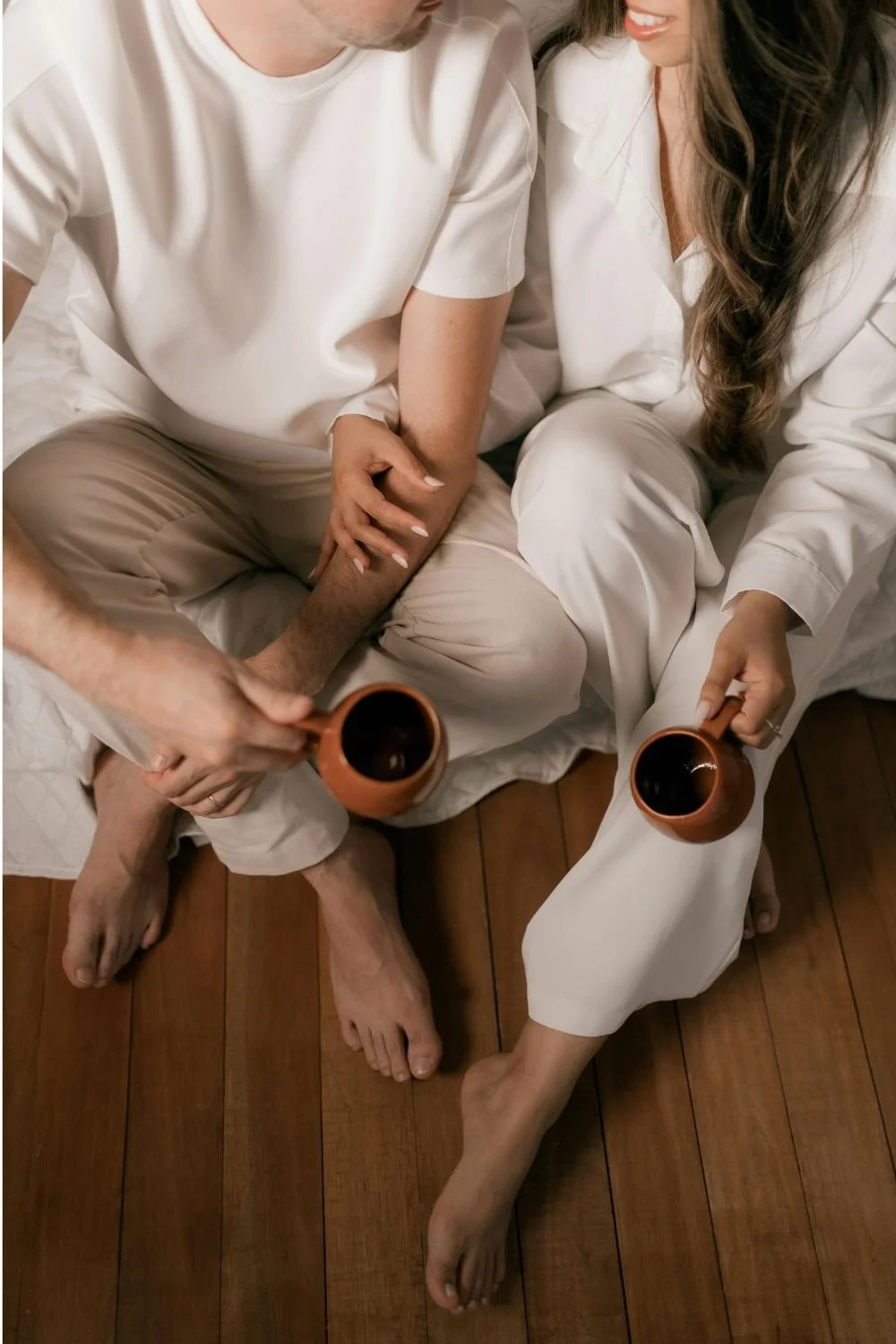 A couple sitting on a bed with wooden floor, holding mugs of coffee, dressed in white pajamas, sharing a quiet moment.