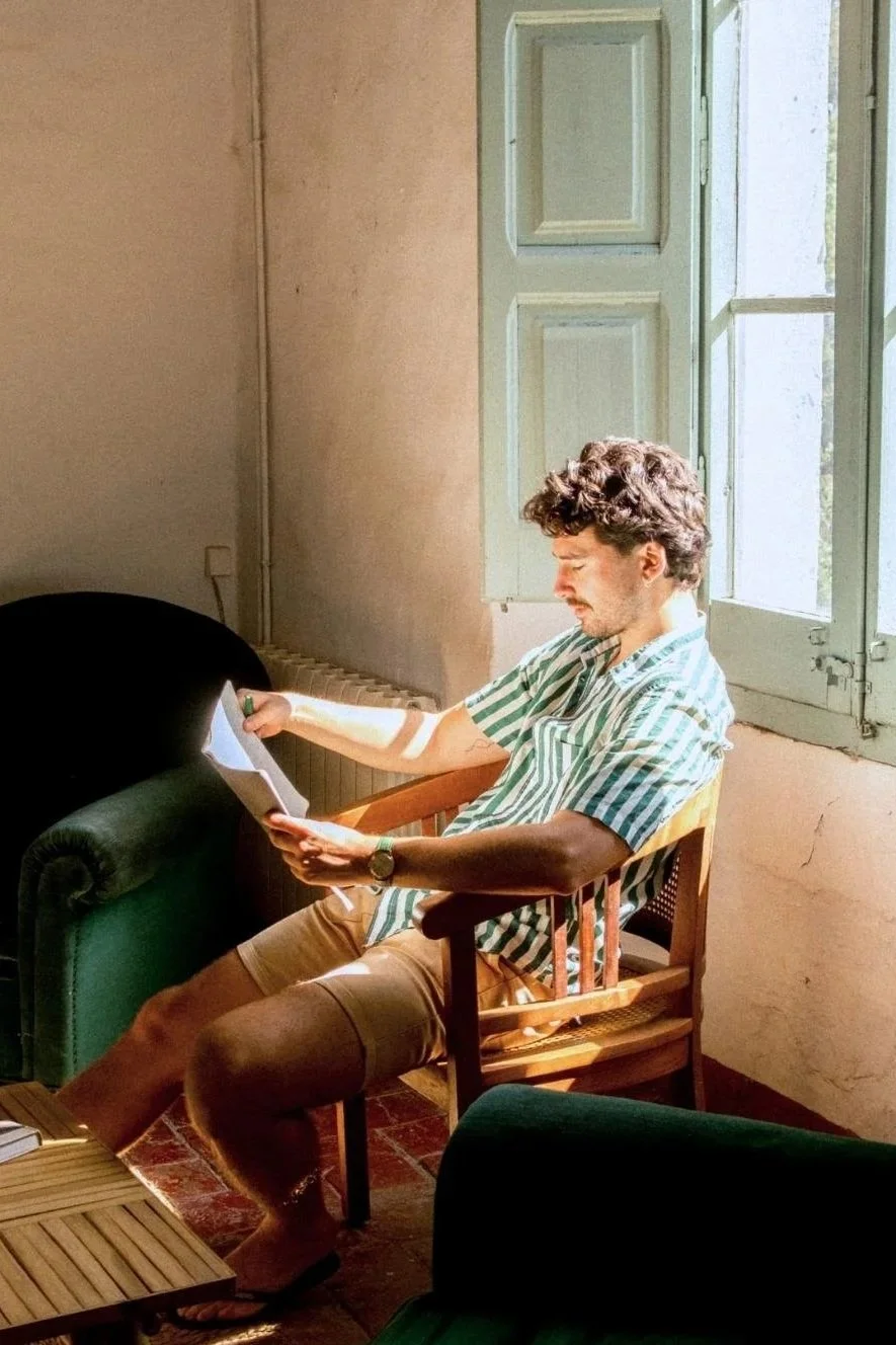 A man with curly hair wearing a striped shirt and shorts, sitting on a wooden chair by a window, reading a book in a sunlit room with a green couch and a brick floor.