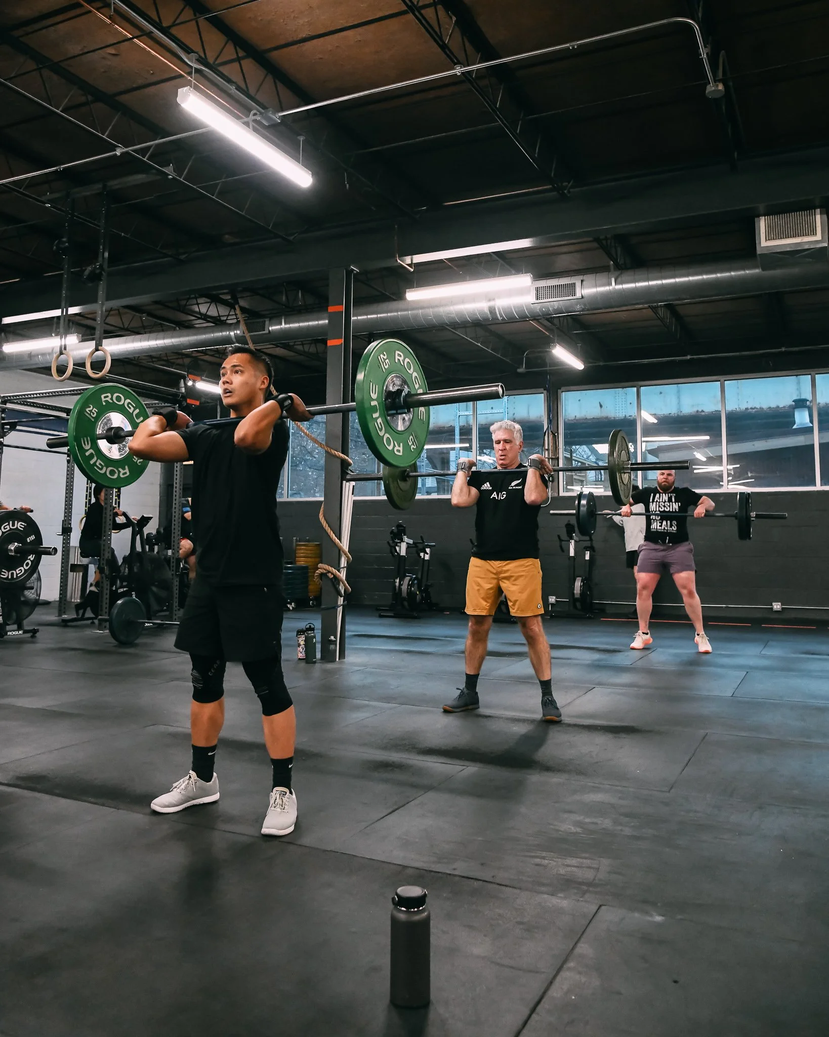 People working out with barbells in a gym, with fitness equipment and gym rings visible in the background.